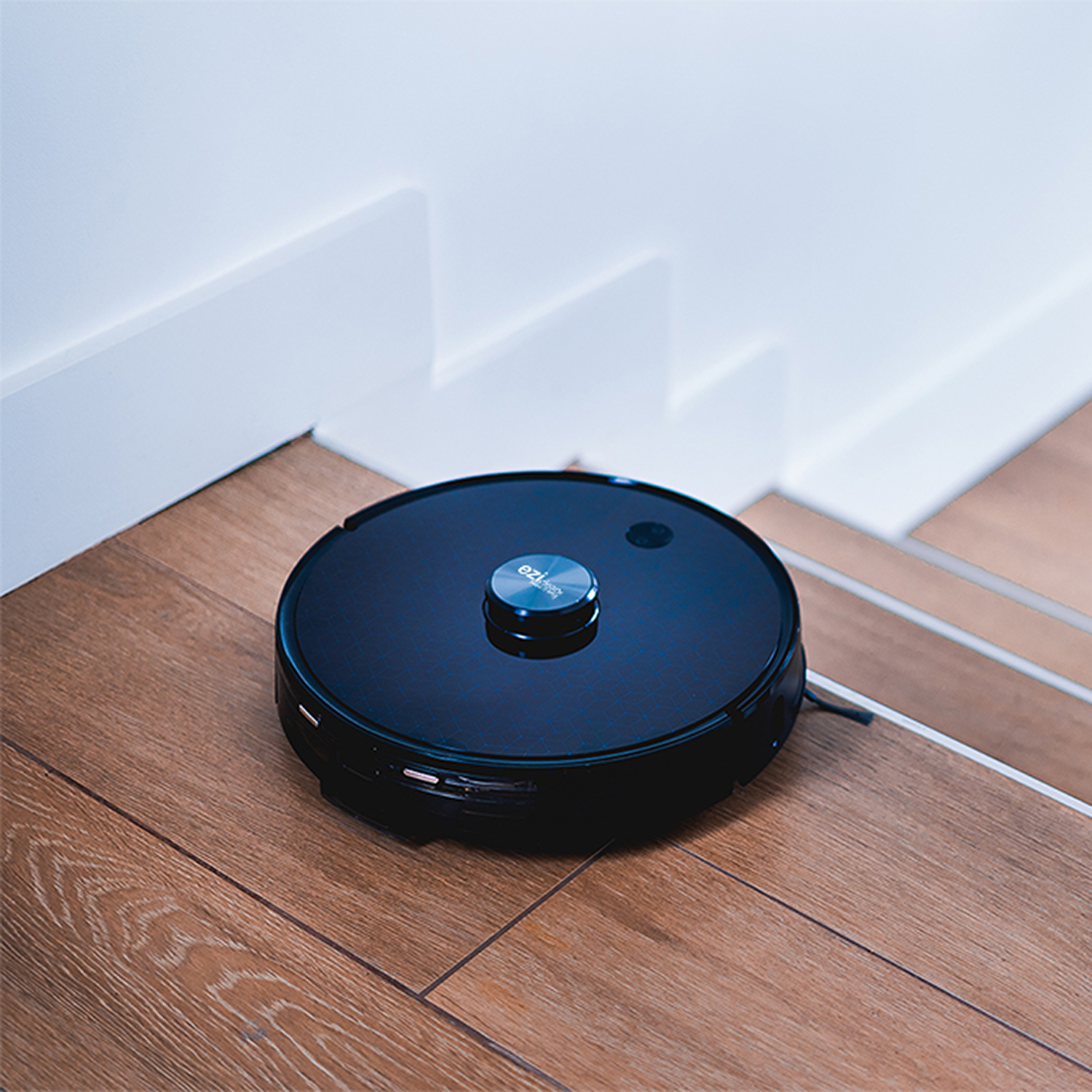 Ein schwarzer, runder Roboter-Staubsauger auf Holztreppen, in der Nähe einer weißen Wand.