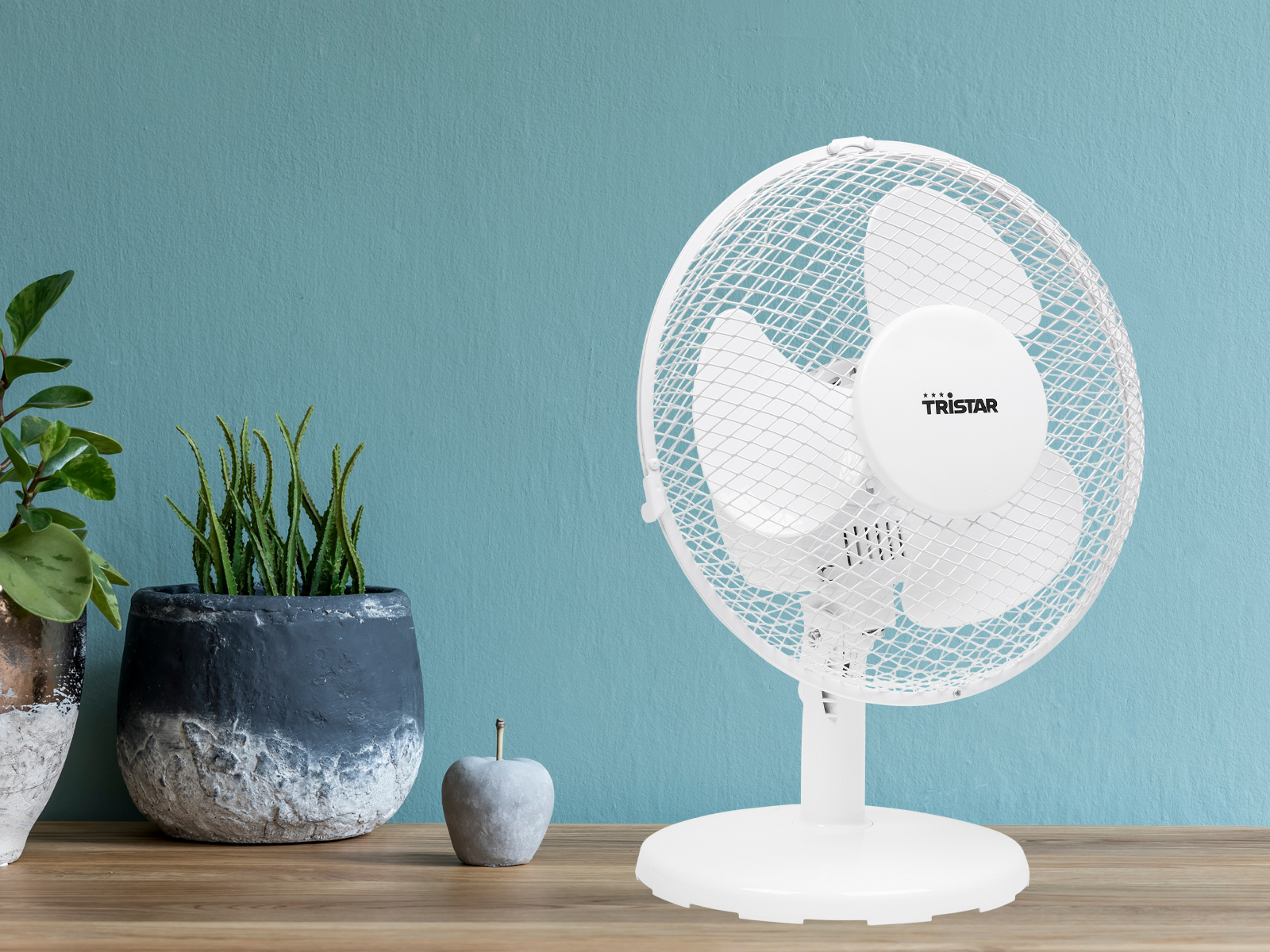 Weißer Ventilator mit Schutzgitter auf einem Ständer, auf Holzoberfläche, mit Pflanzen und Apfel.