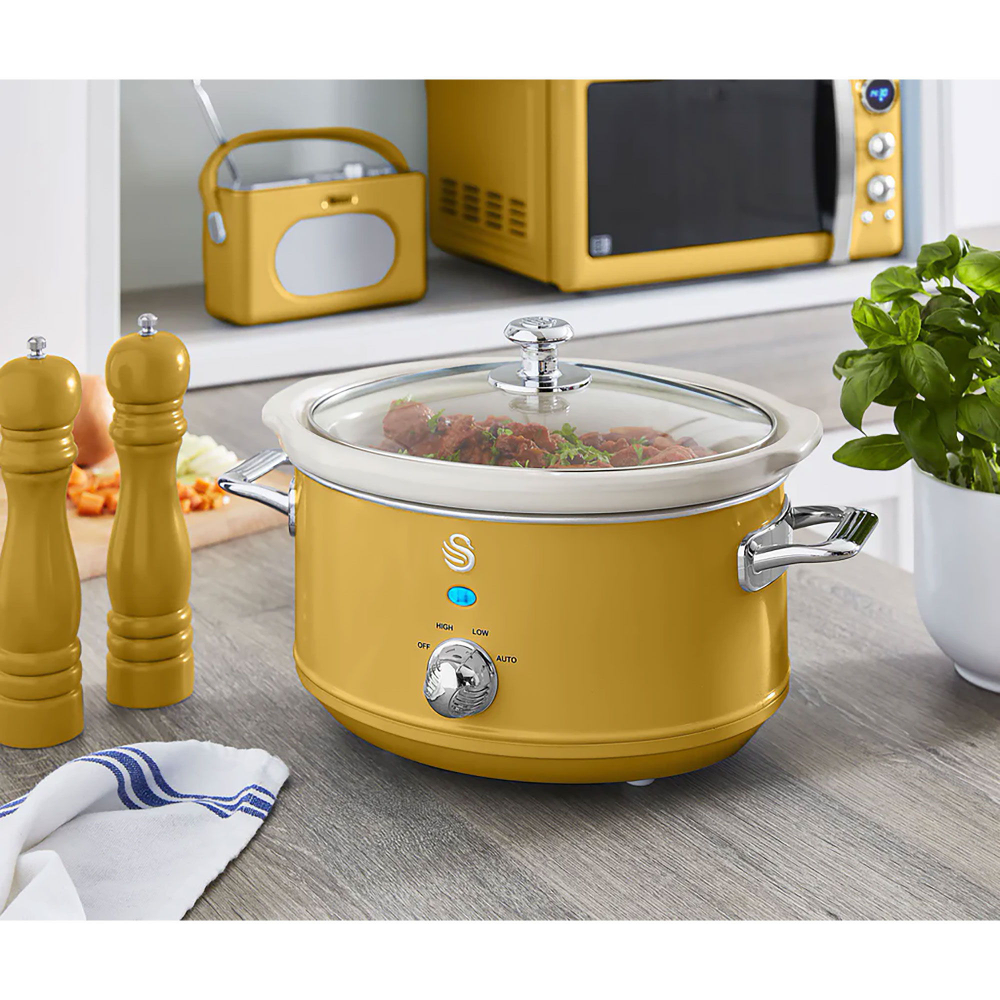 Ein senfgelber Slow Cooker mit einem durchsichtigen Deckel steht auf einer grauen Theke. Hinten steht eine gelbe Mikrowelle.