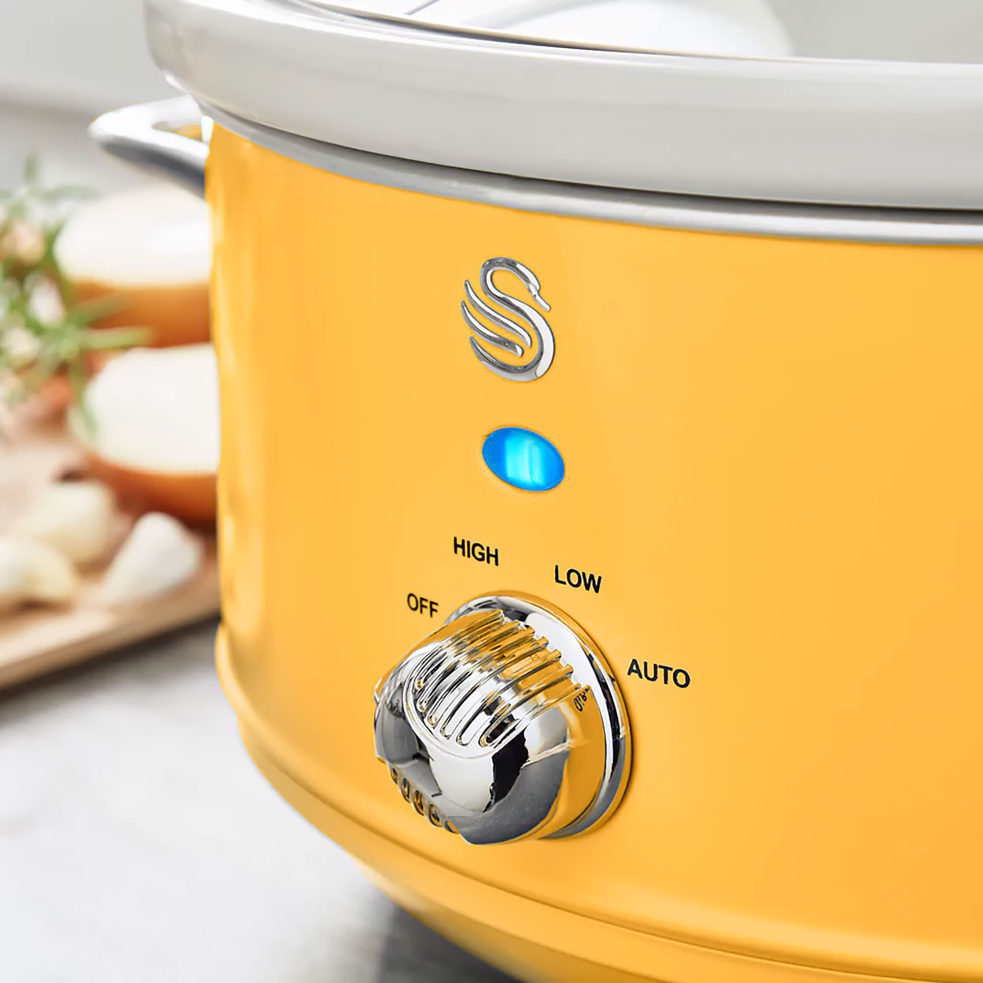 Gelber Slow Cooker mit Chrom-Drehrad und blauem Licht. Zwiebeln und Knoblauch im Hintergrund.