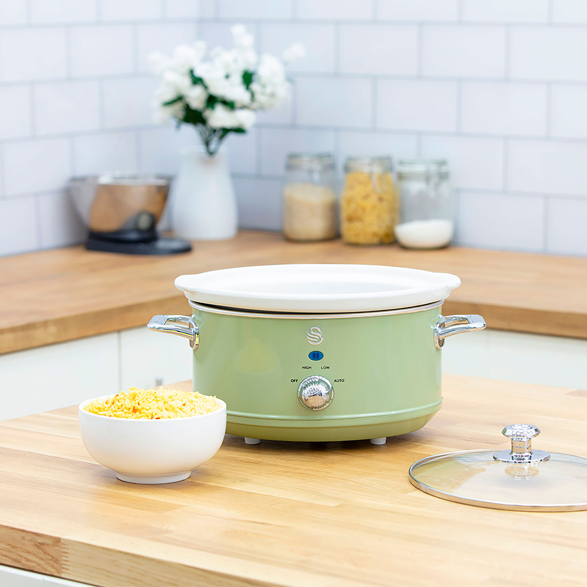 Grüner Slow Cooker auf einer Holztheke, mit einer Schüssel Essen und einem Glasdeckel.