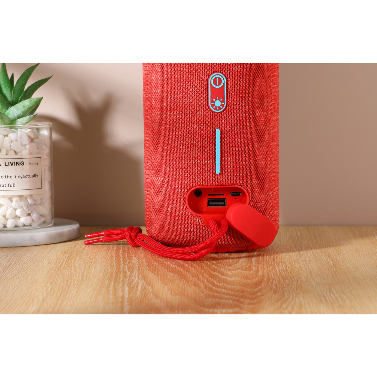 Altavoz cilíndrico rojo con botones y puertos. Tiene una correa roja. Una planta pequeña en el fondo.