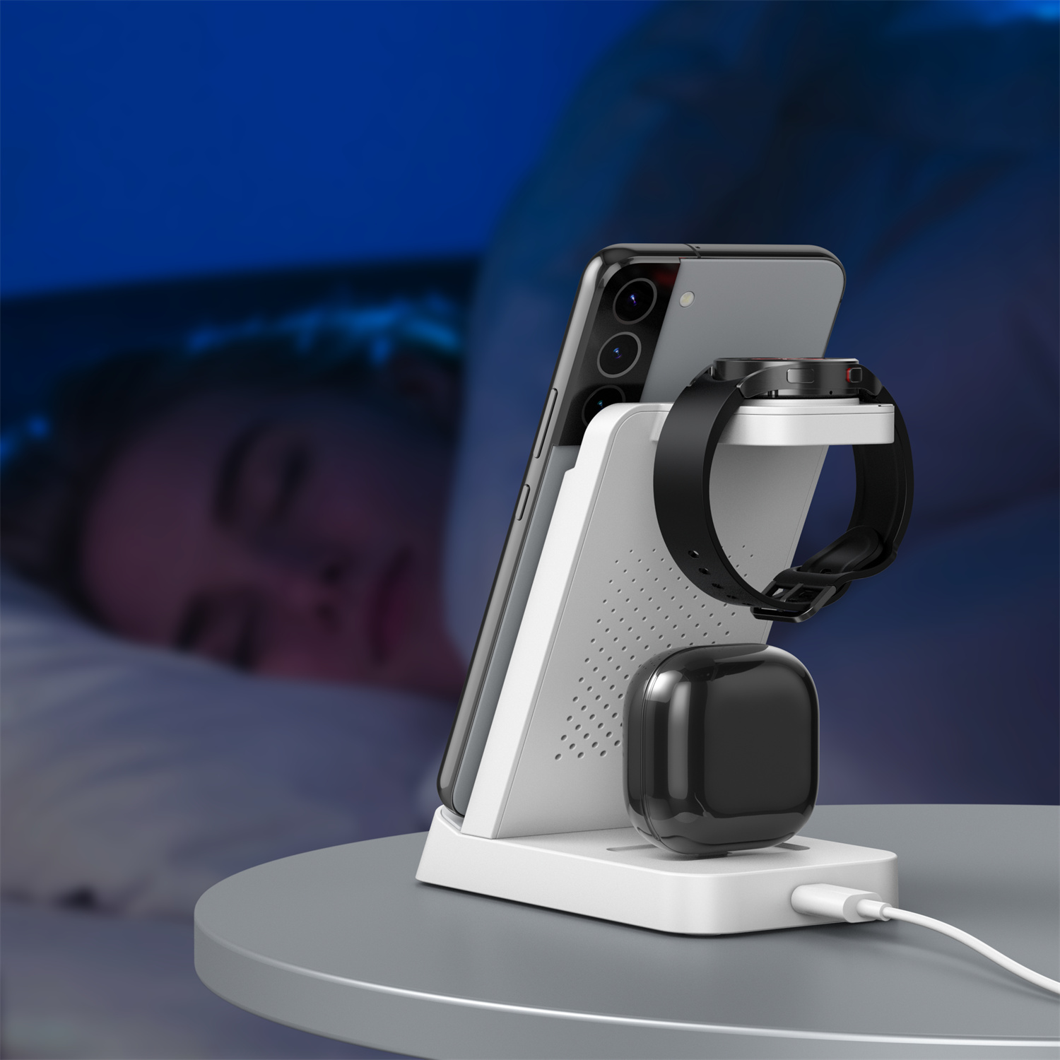 Stacja ładująca z telefonem, zegarkiem i słuchawkami. Osoba śpi w tle, oświetlona niebieskim światłem.