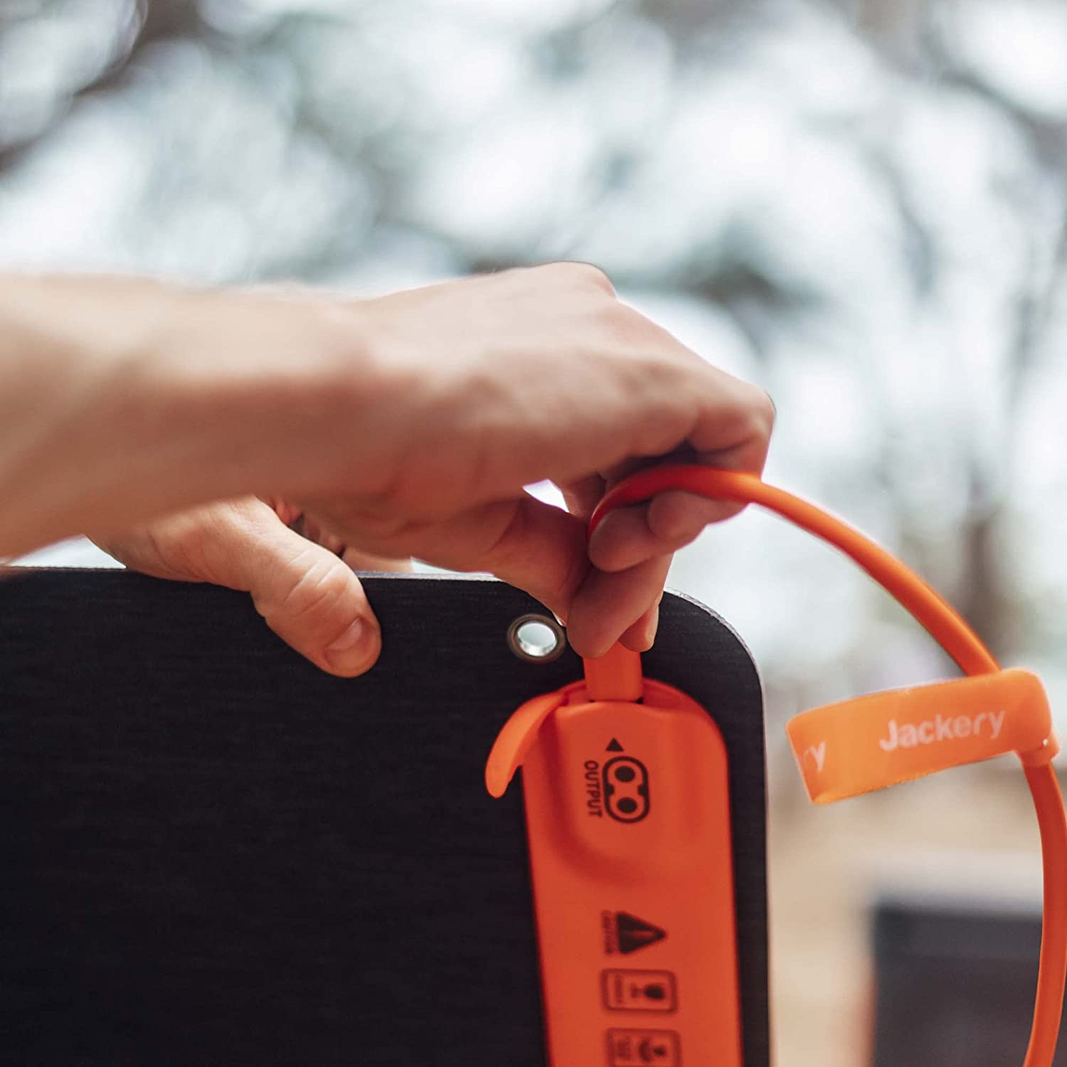 Hand die een oranje oplaadkabel aansluit op een zwarte Jackery powerbank. Vage achtergrond.