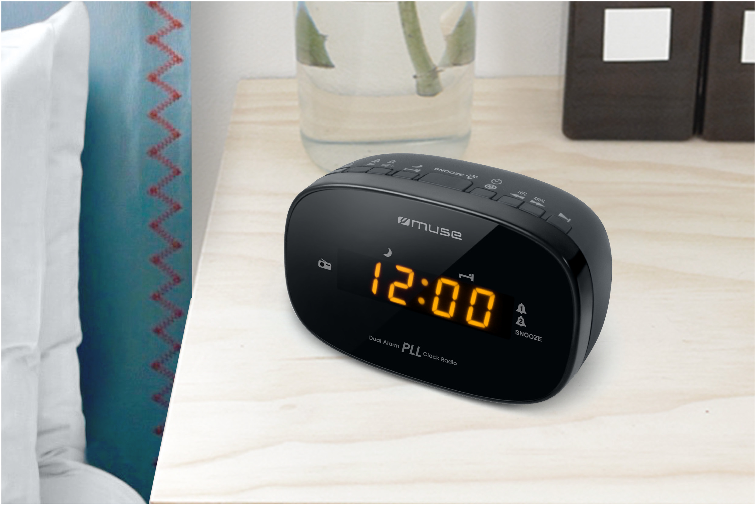 Black clock radio showing 12:00 on a wooden table, with a glass vase and pillow partially visible.