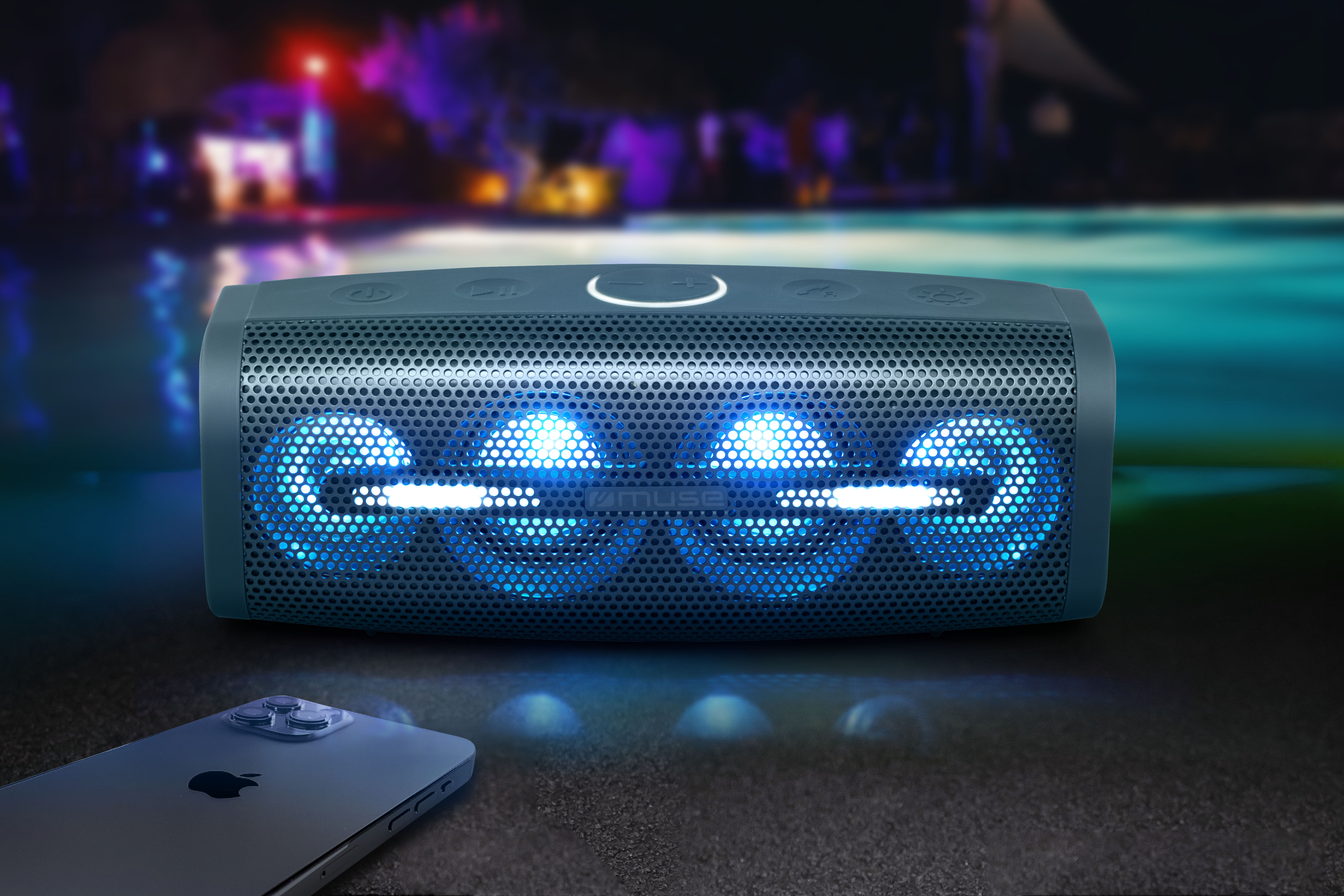 Czarny głośnik z niebieskimi światłami i iPhone na piaszczystej powierzchni, z wodą i rozmytym tłem.