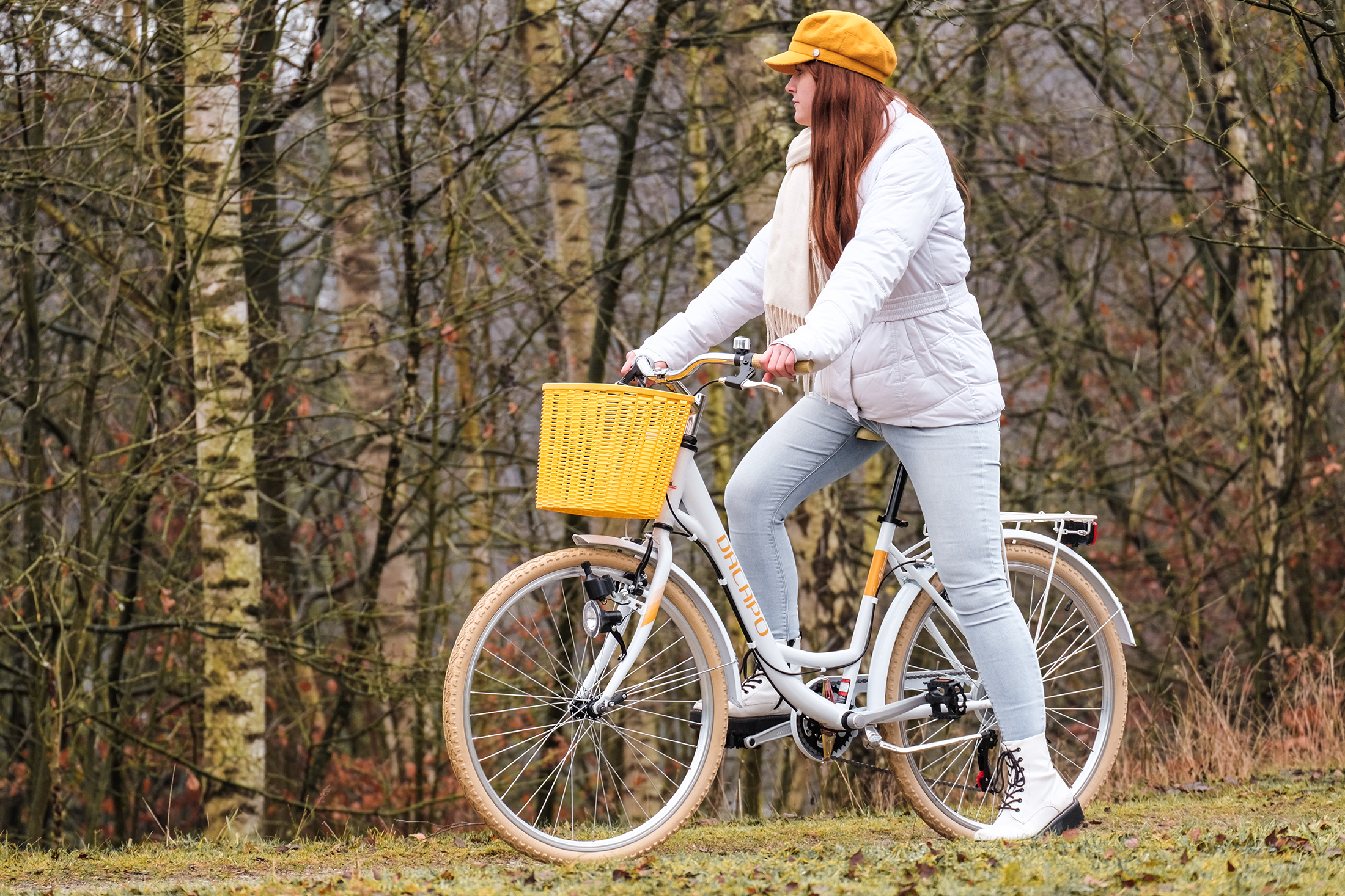 Frau fährt mit einem weißen Fahrrad mit gelbem Korb. Sie trägt einen gelben Hut und einen weißen Mantel.