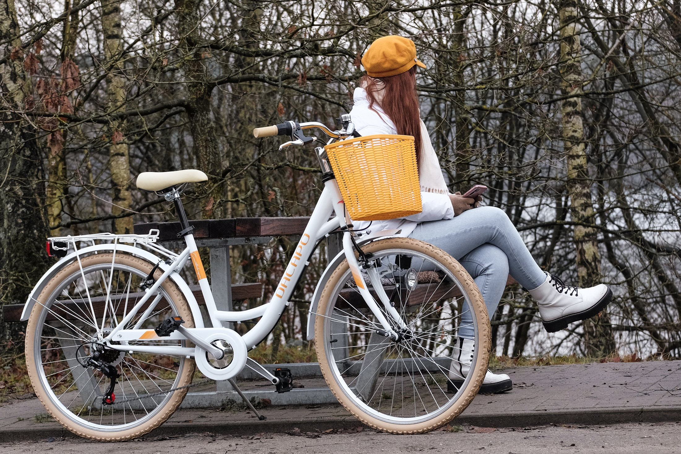 Frau auf weißem Fahrrad mit Korb, sitzt auf einer Bank. Herbstfarben, kahle Bäume und See im Hintergrund.