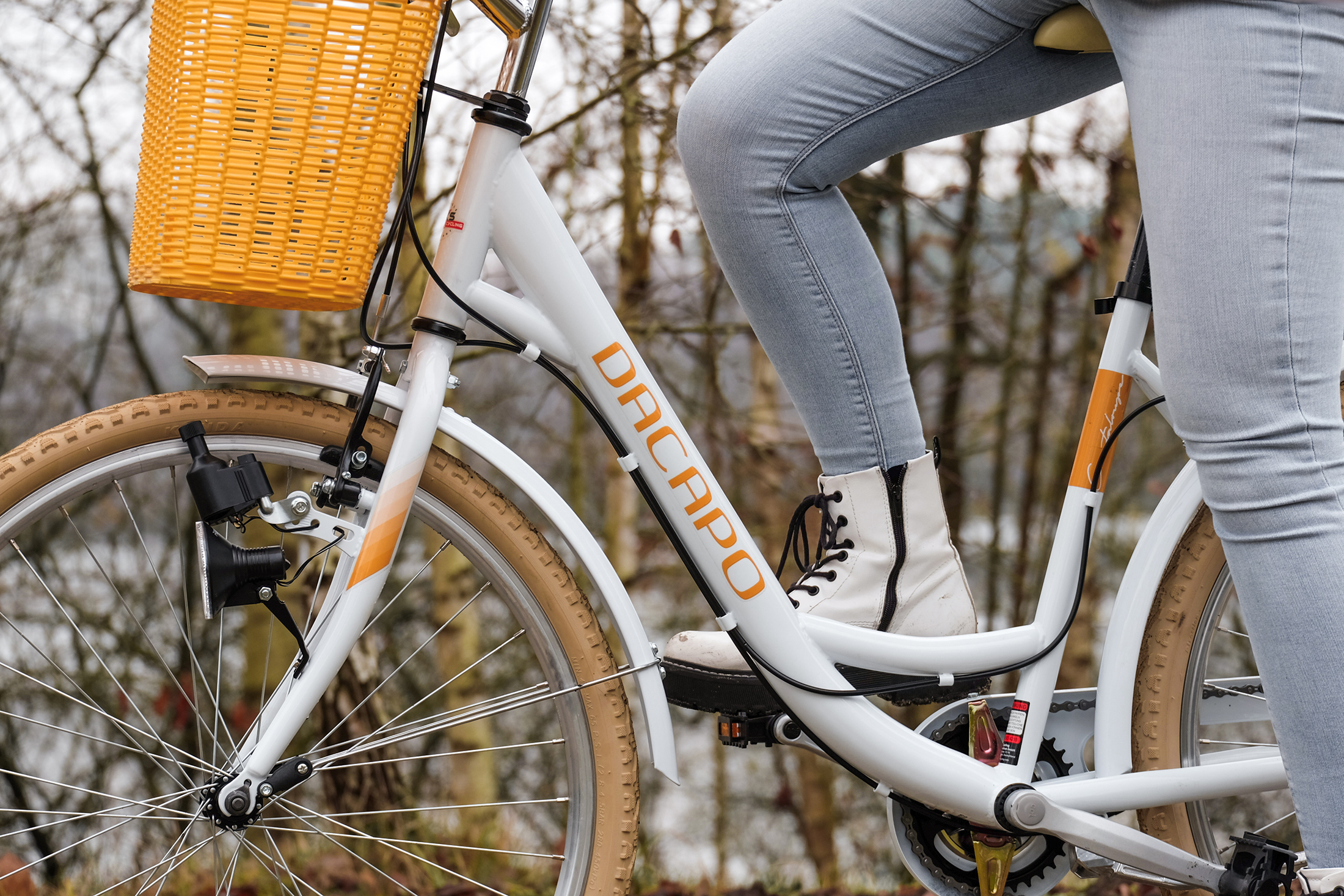 Eine Frau fährt ein weißes Fahrrad mit orangefarbenem Korb. Bäume sind im Hintergrund.