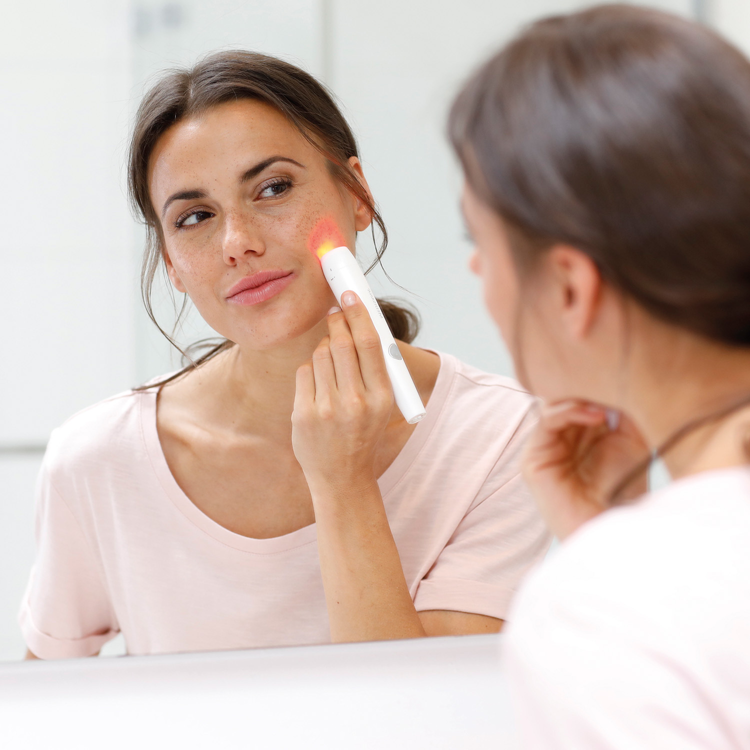 Woman looking in mirror uses a skin treatment device. Red light on skin.
