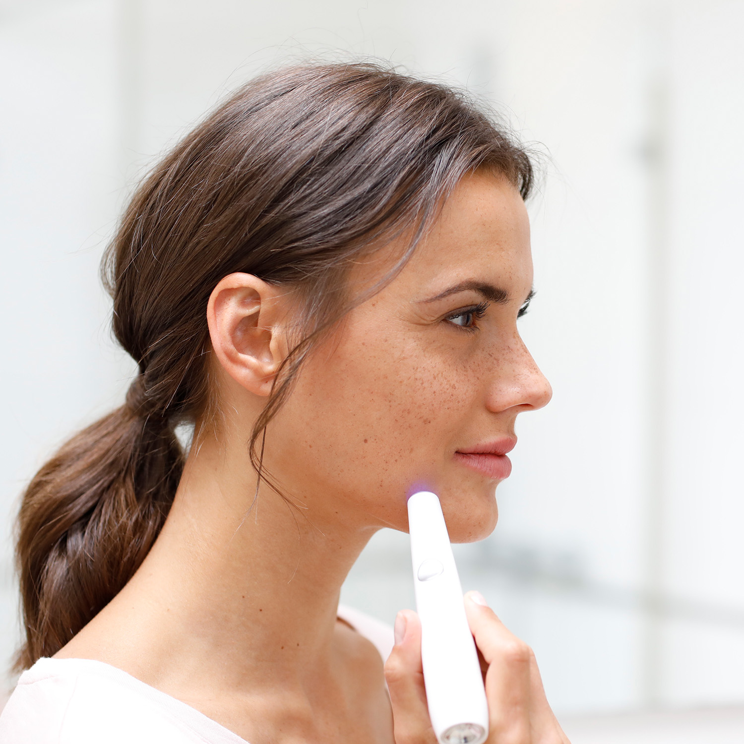 Woman using a white handheld device on her cheek.  The device emits a purple light.