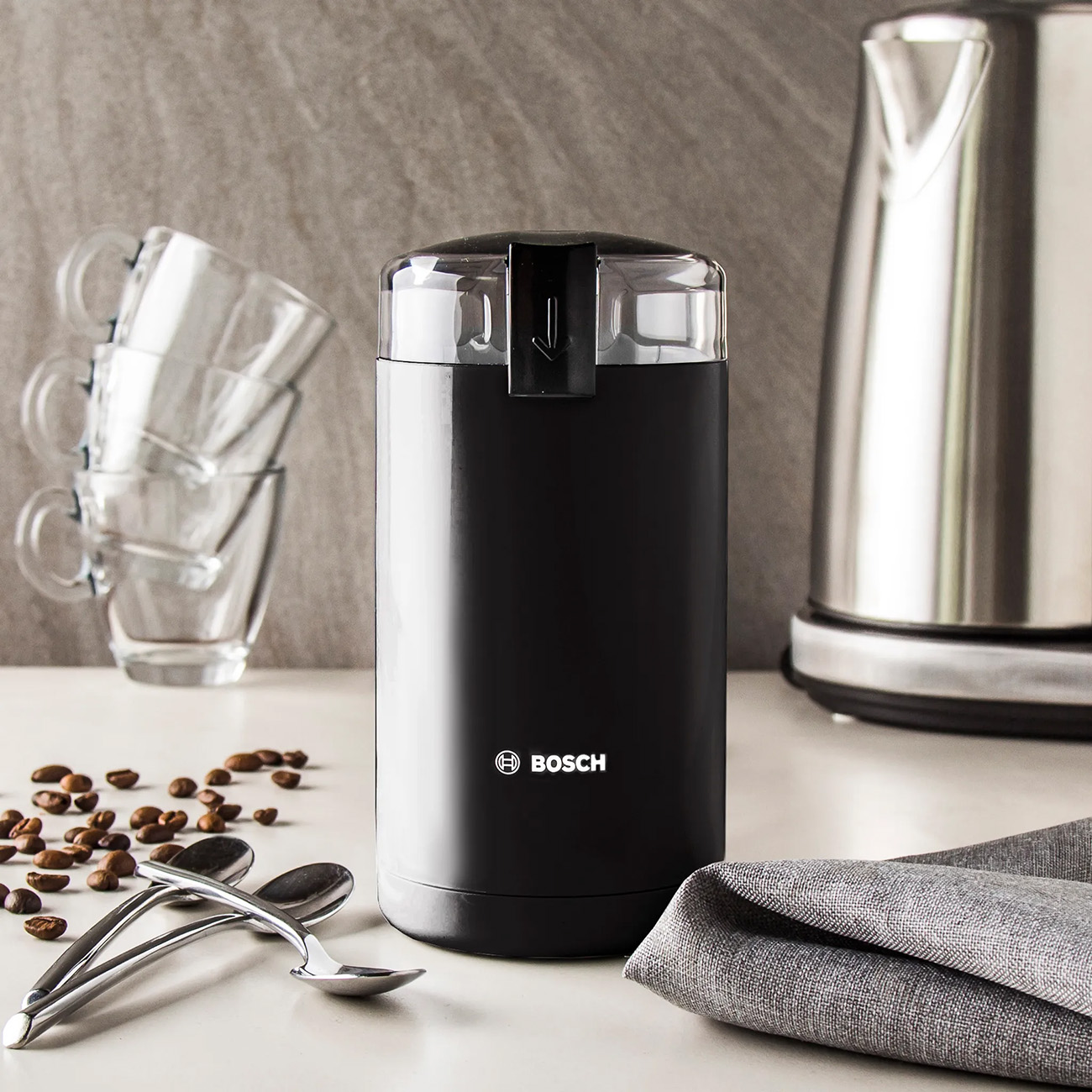 Black coffee grinder with a clear top, coffee beans, spoons, and a kettle.