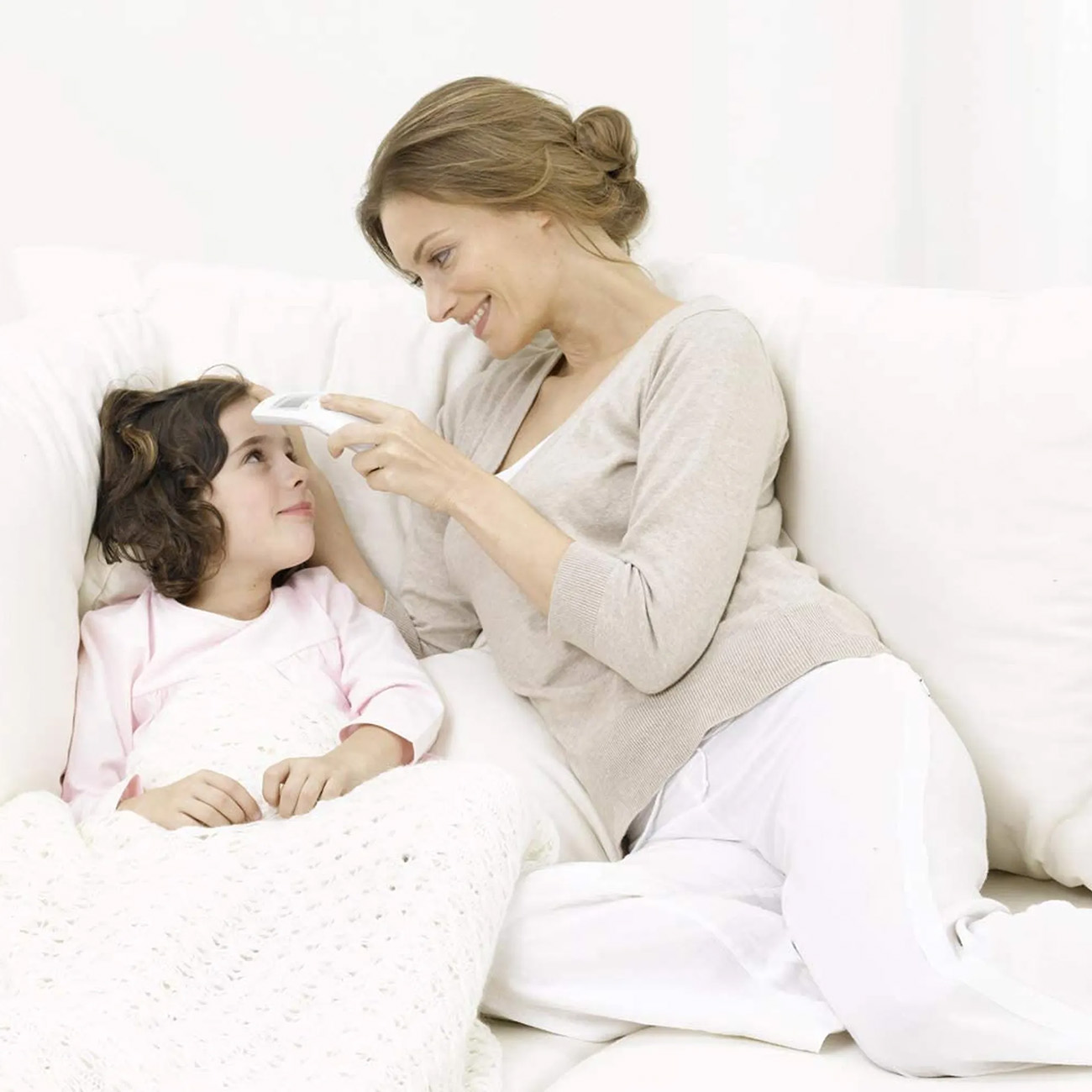 A woman taking a child's temperature with a digital thermometer. Both are on a white couch.
