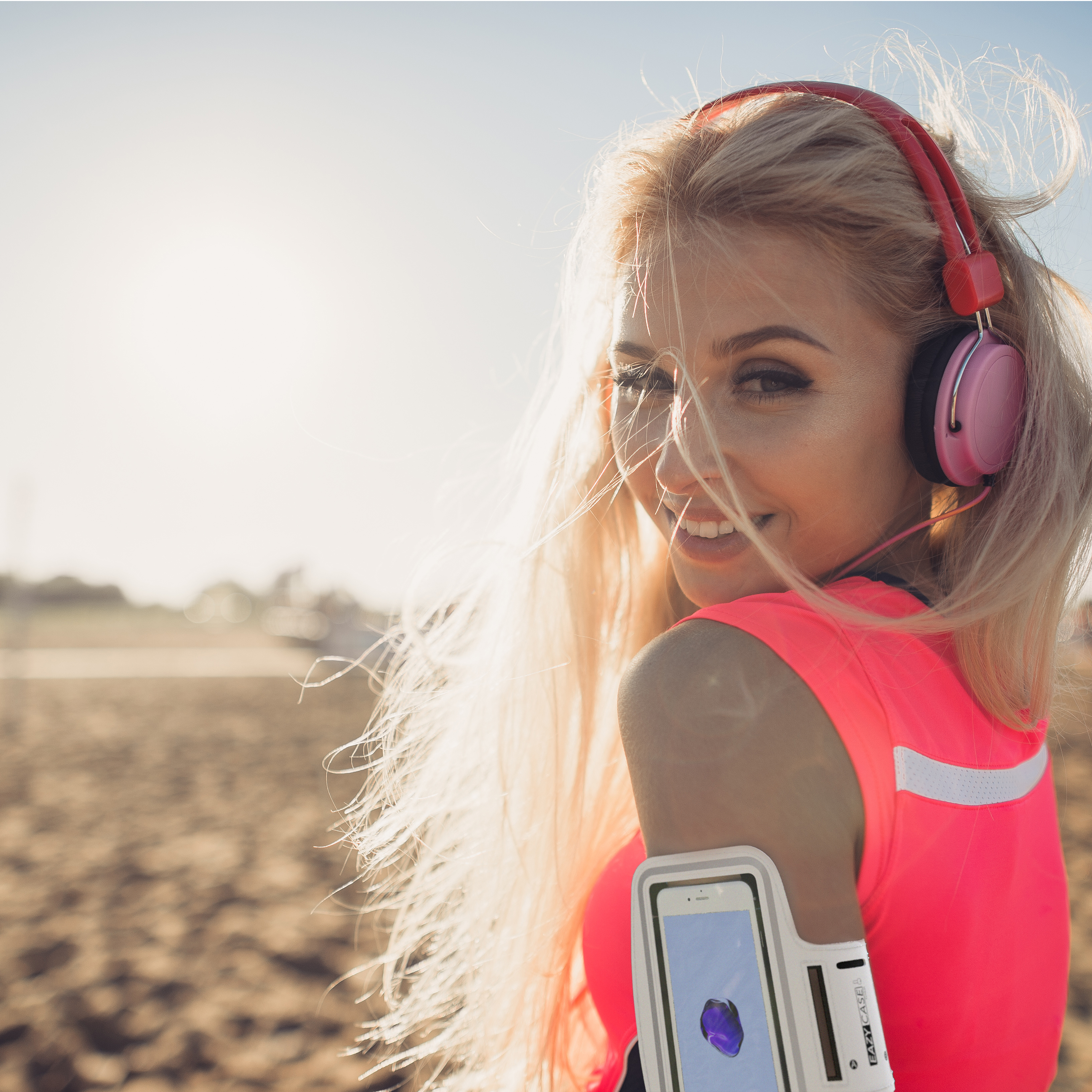 Frau mit blonden Haaren, lächelnd, mit pinkfarbenen Kopfhörern und Shirt, Armband mit Telefon.