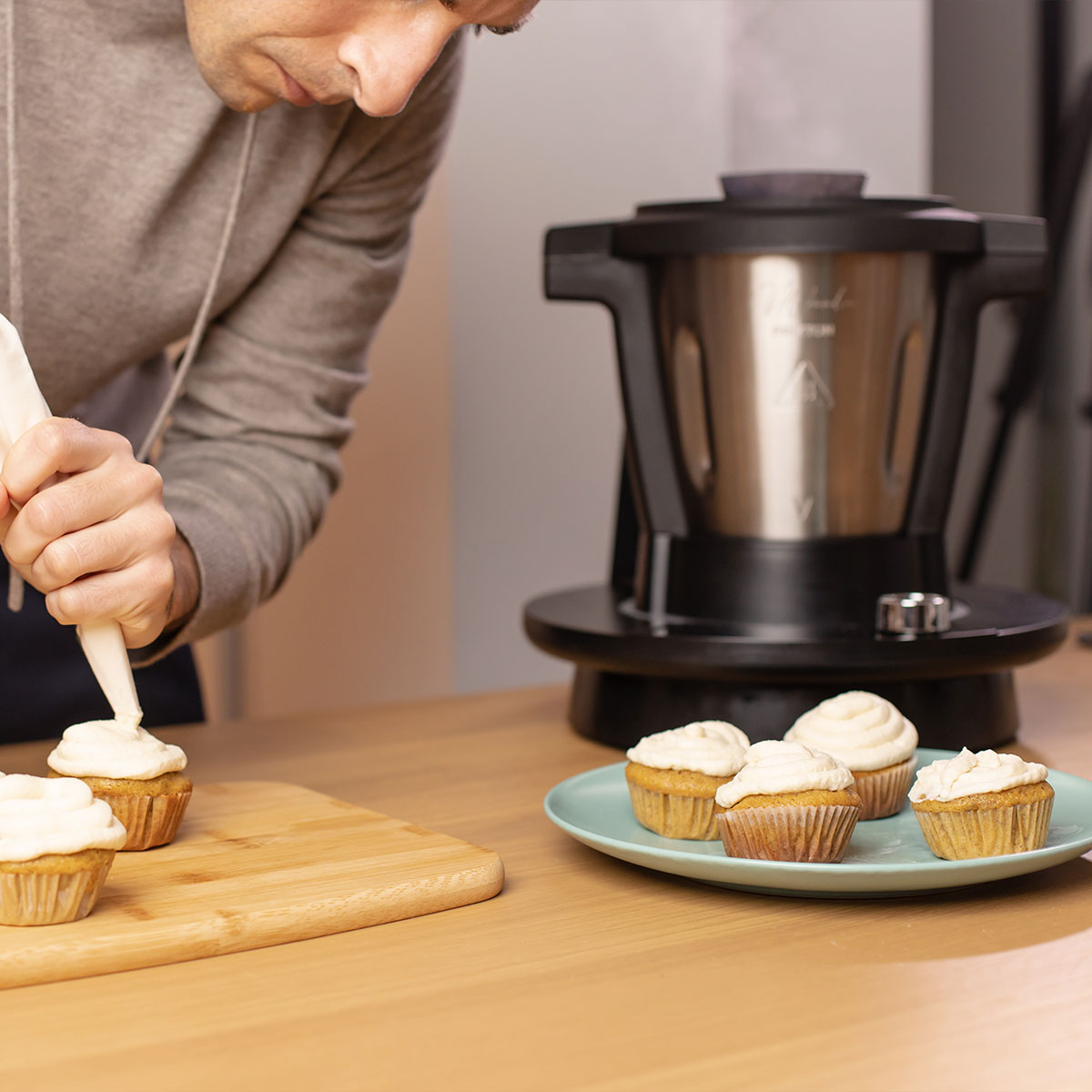 Osoba dekoruje babeczki lukrem za pomocą rękawa cukierniczego na drewnianej desce.