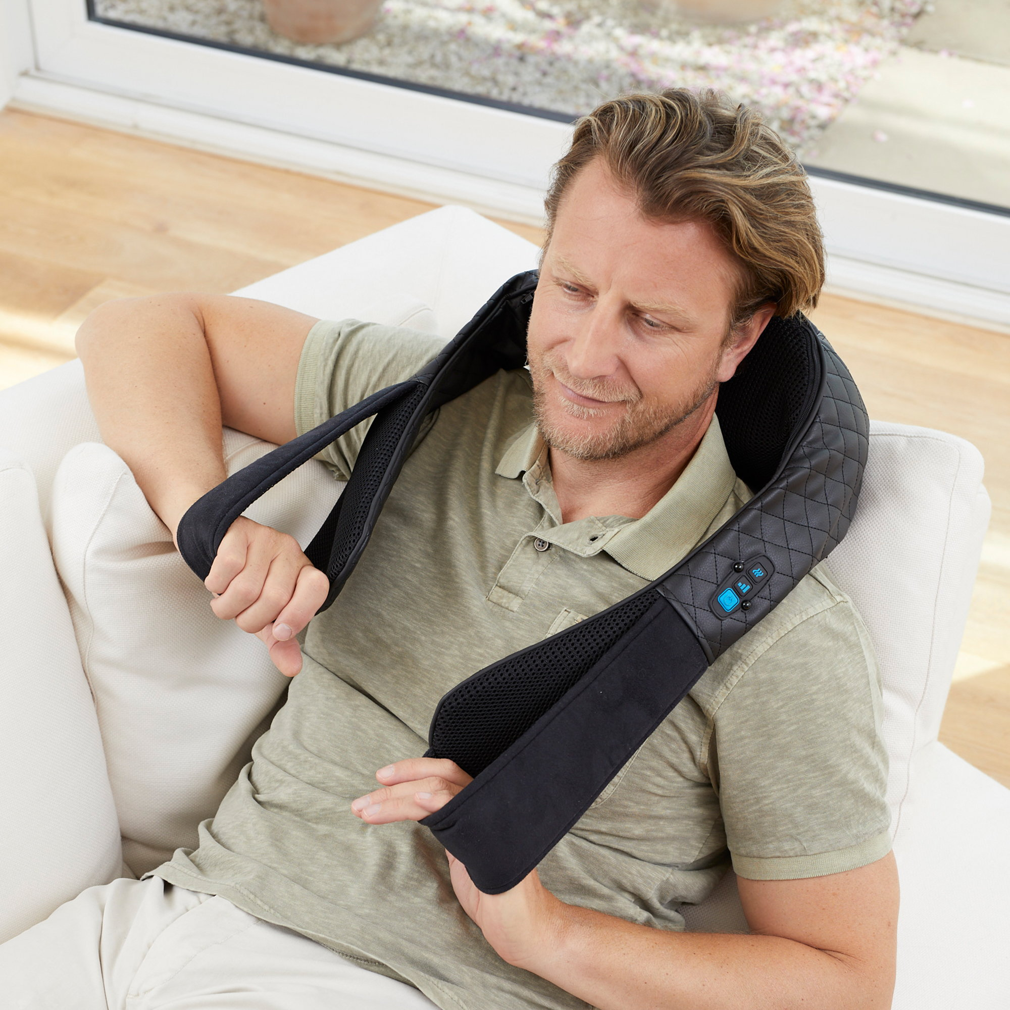 Mężczyzna noszący czarny masażer szyi na białej kanapie i dotykający dłoni.