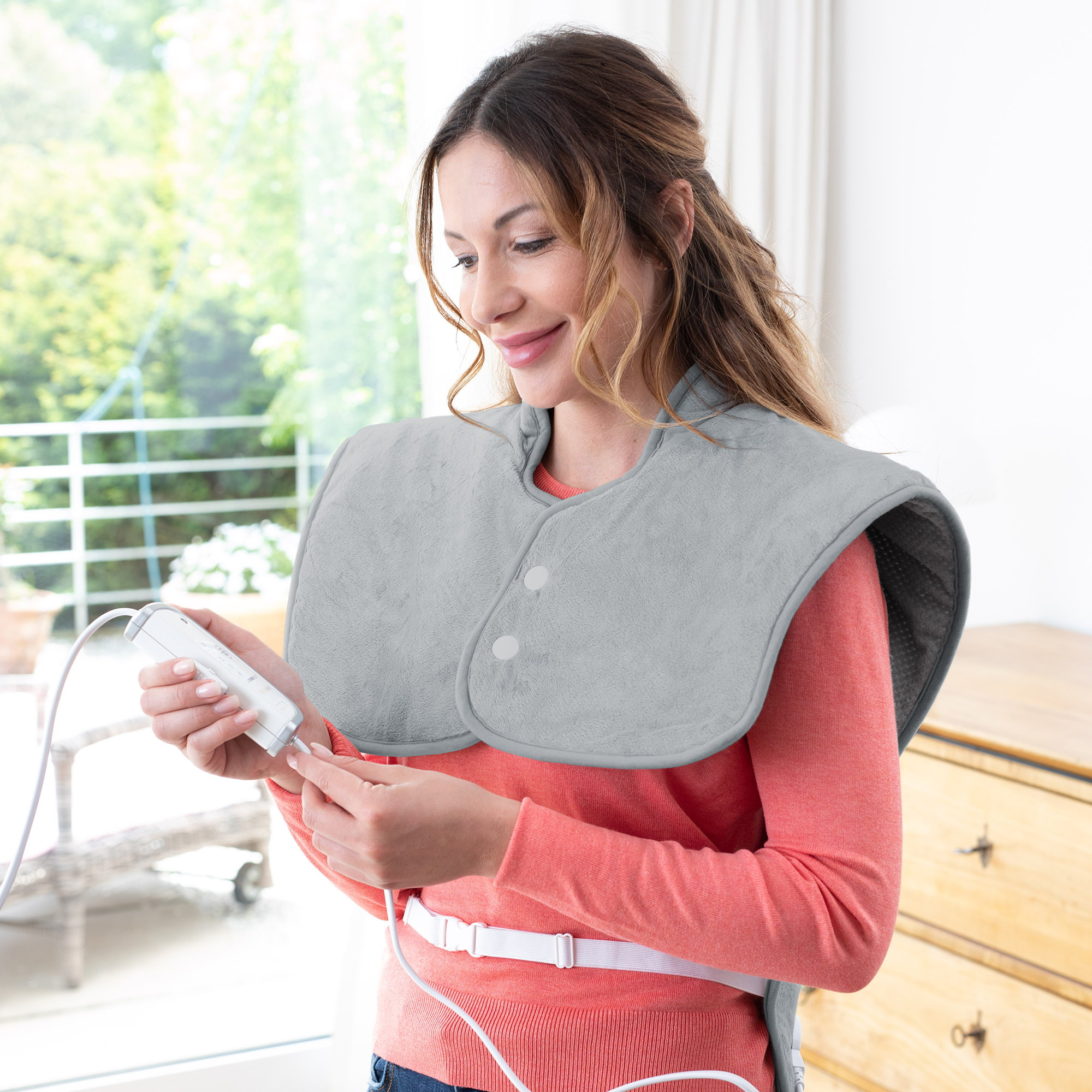 Kobieta trzyma urządzenie podłączone do szarej poduszki grzewczej. Uśmiecha się w pokoju.