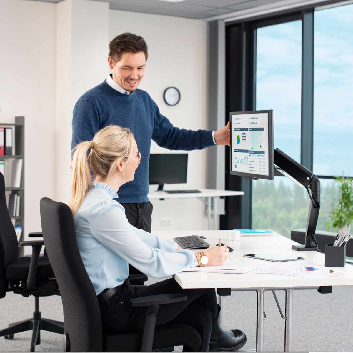 Dos personas en una oficina; una señala un monitor con gráfico, la otra lo mira.