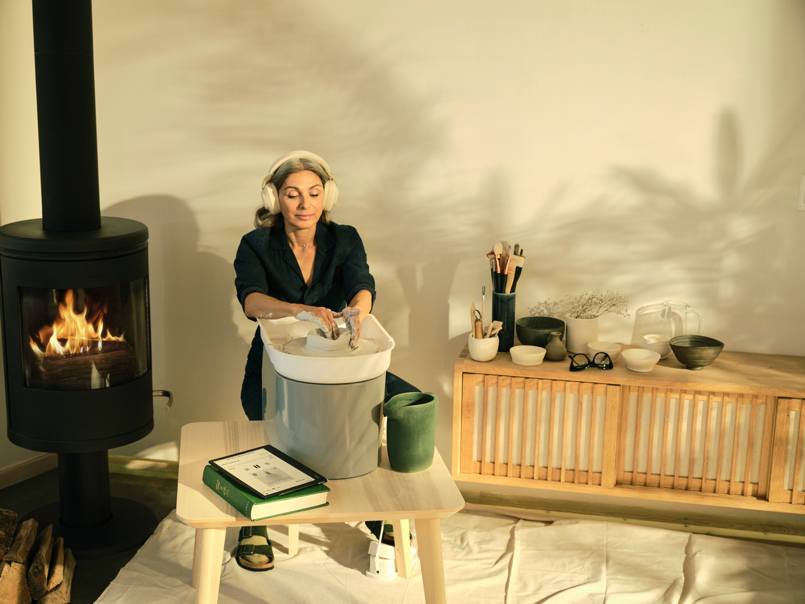 Kobieta przy kole garncarskim, kominek i drewniana szafka w pokoju.