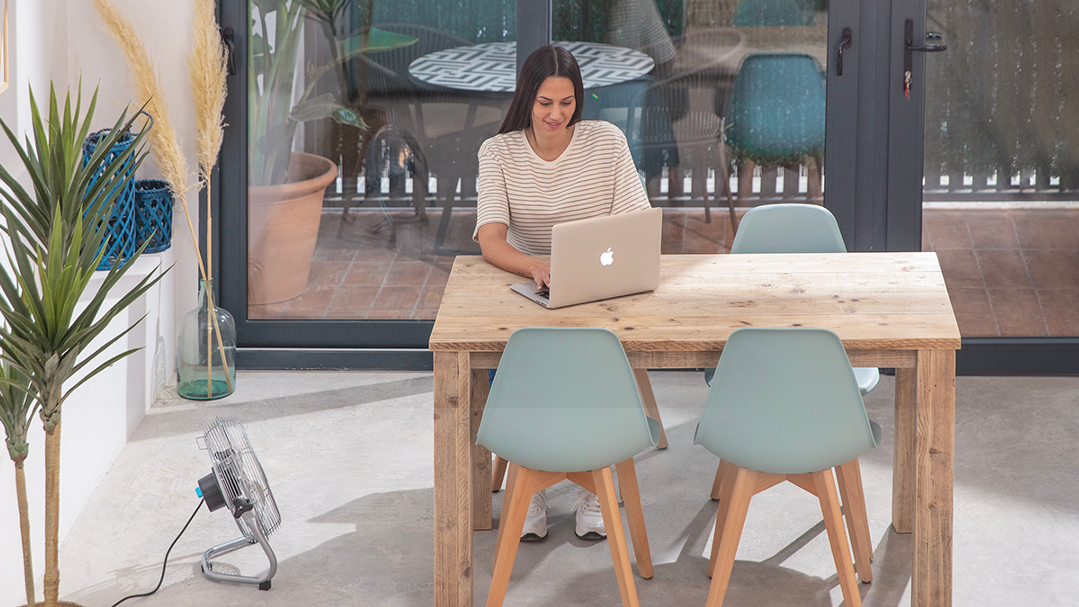 Kobieta pracuje na laptopie przy drewnianym stole, z krzesłami i wentylatorem. Scena jest na zewnątrz.