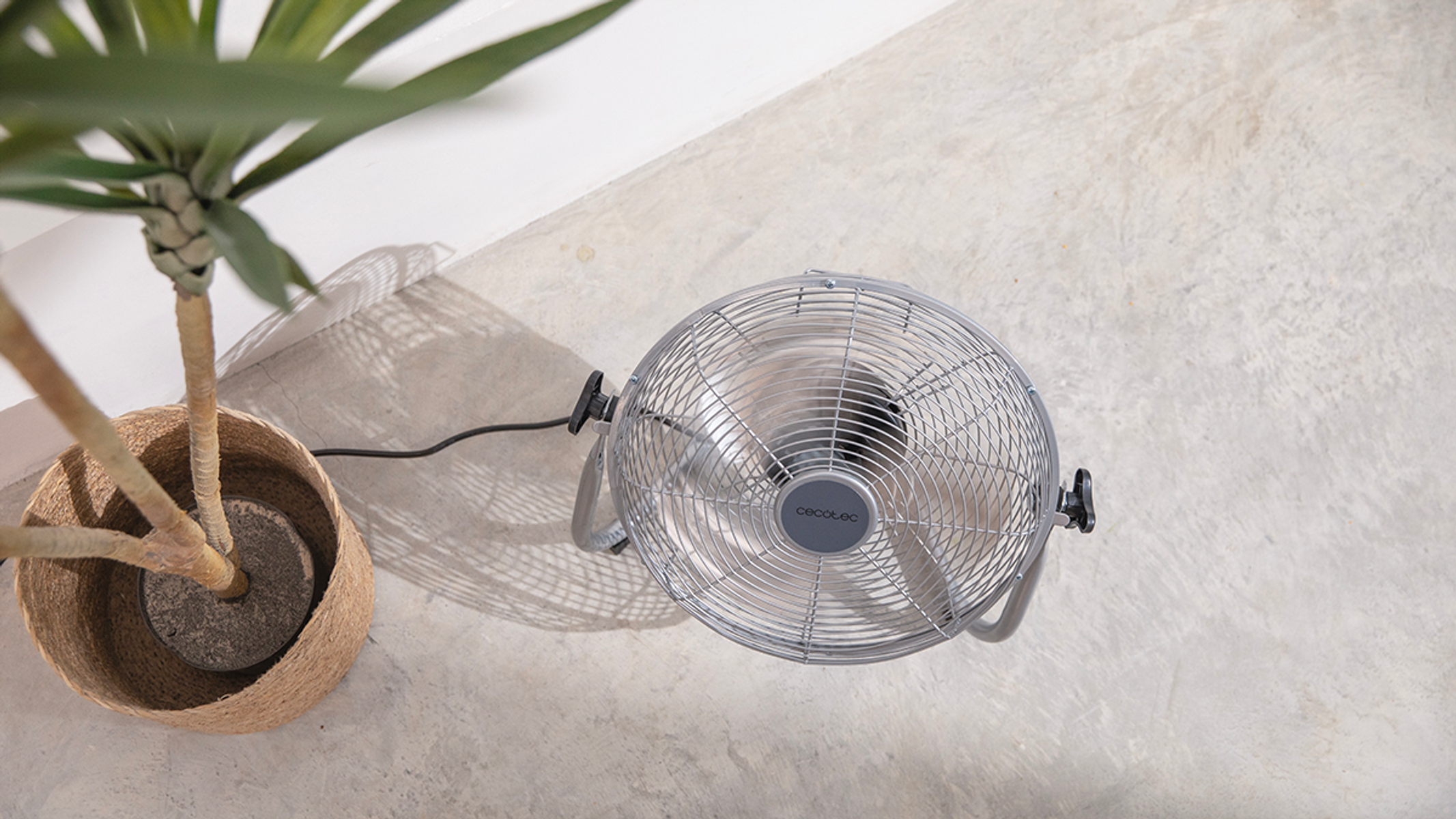Szary wentylator na betonowej podłodze obok rośliny w wiklinowym koszu.