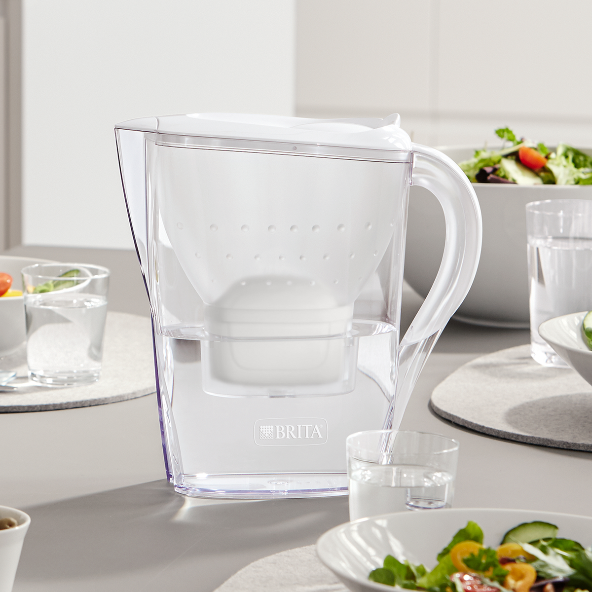 Ein klarer Brita Wasserfilter-Krug auf einem Tisch mit Wasser und Salat. Er ist mit Wasser gefüllt.