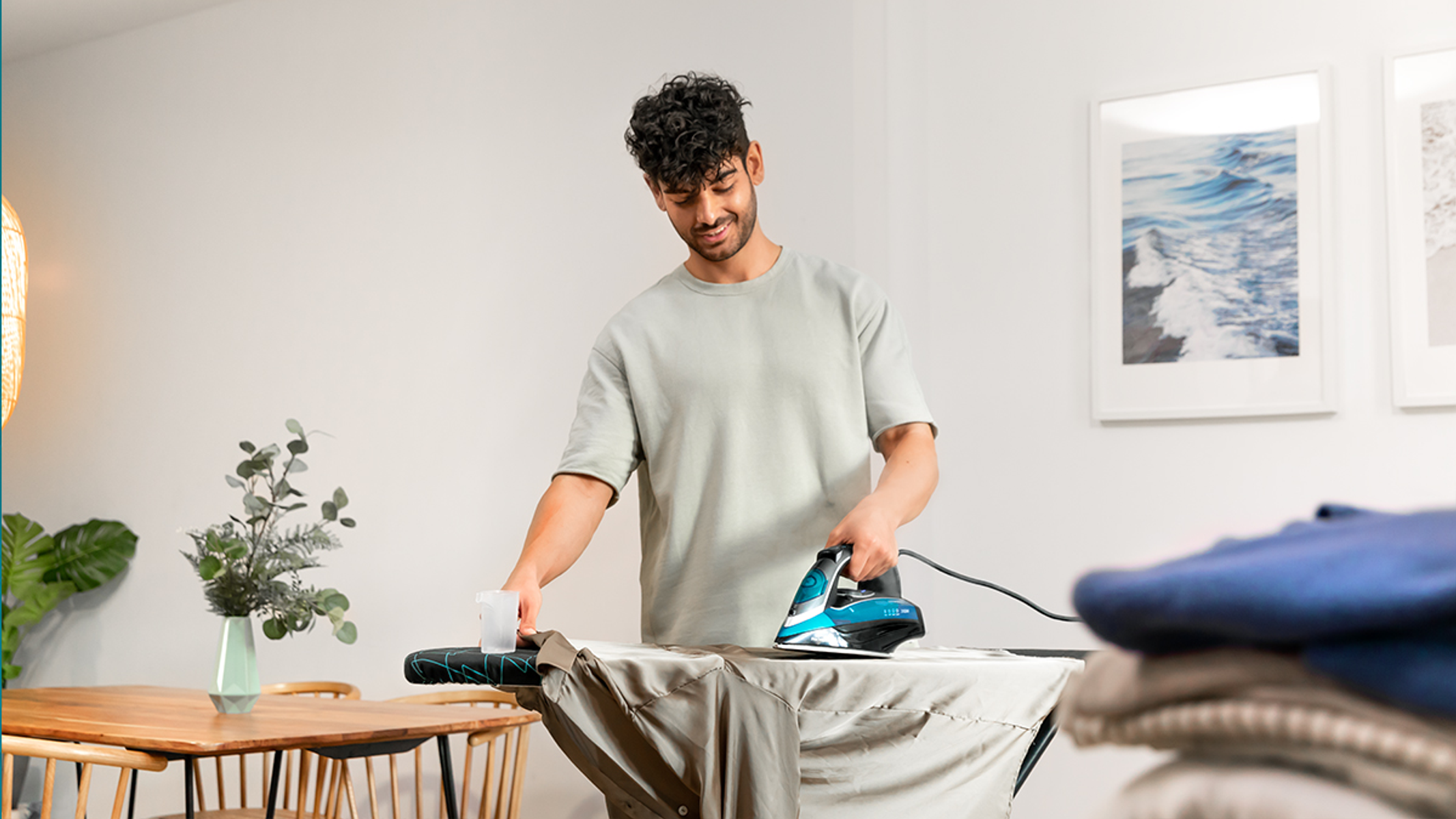 Un hombre planchando una camisa en una tabla de planchar. Hay ropa cerca.