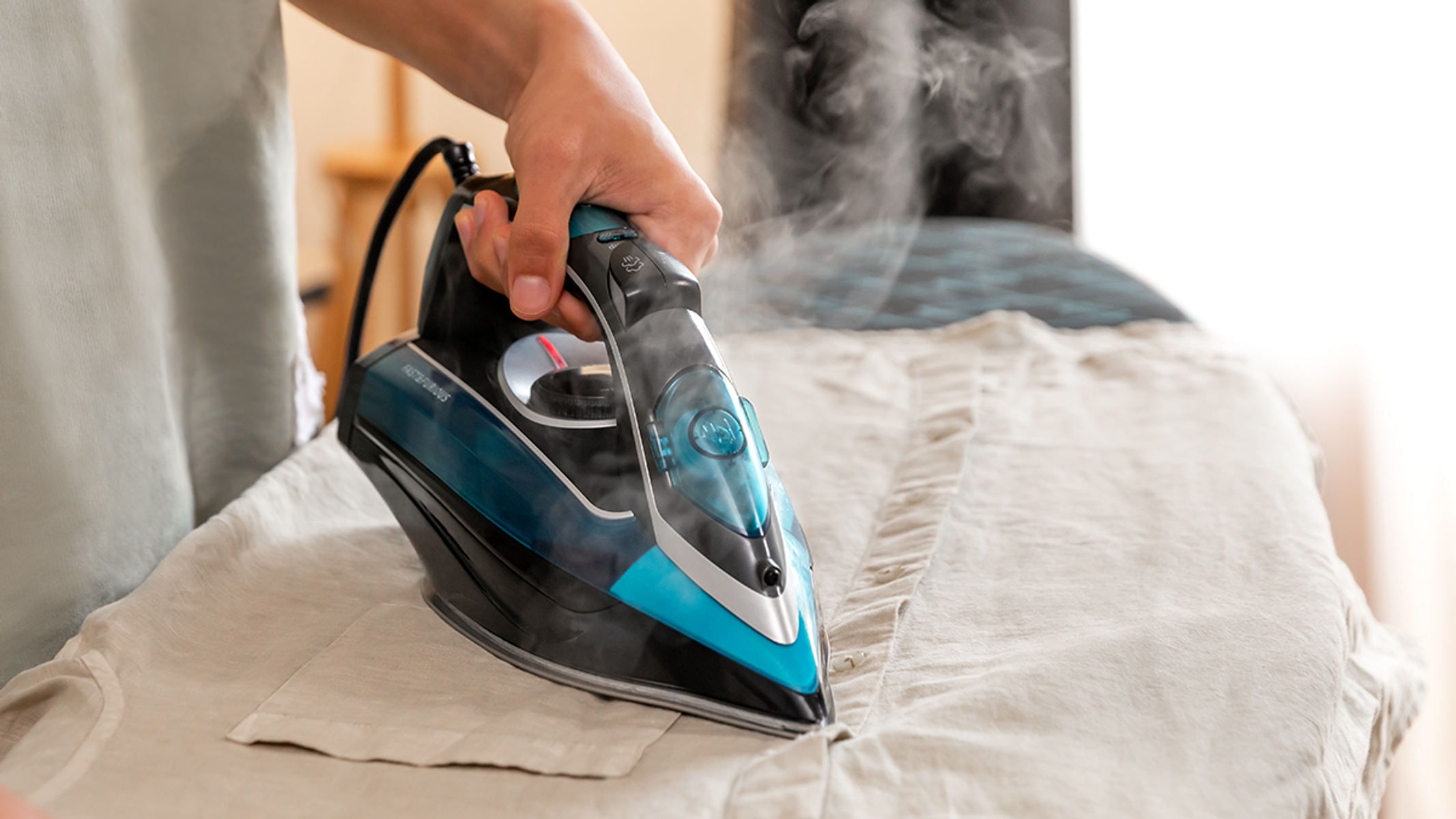 Una plancha vaporizando una camisa beige sobre una tabla de planchar, sostenida por una mano.