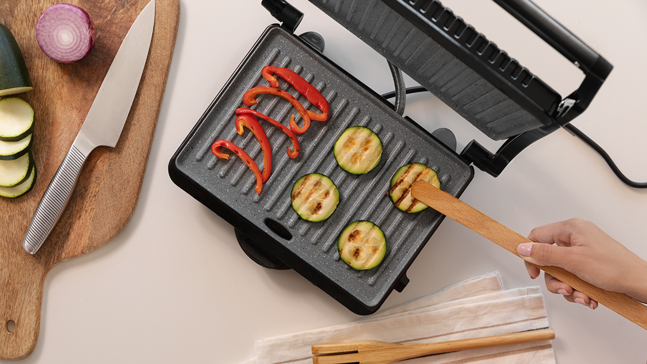 Grill z grillowaną cukinią i czerwoną papryką. Obok nóż i plasterki cukinii.