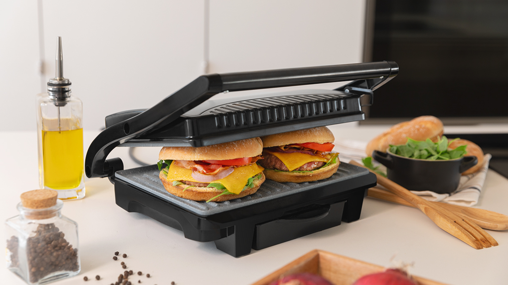 Grill z dwoma burgerami, serem i boczkiem. Butelka z olejem i składniki w tle.