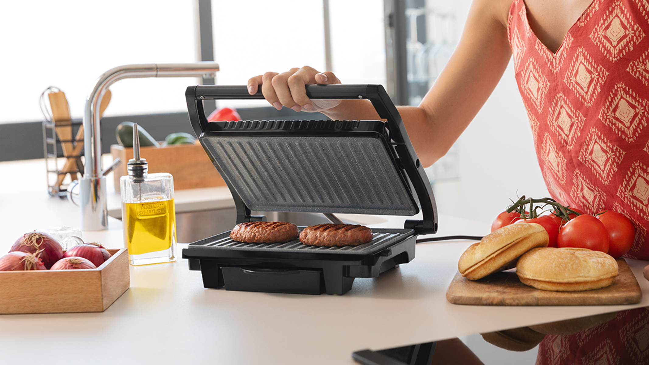 Kobieta używa grilla do smażenia dwóch burgerów. Pomidory, bułki, cebula i olej są na blacie.