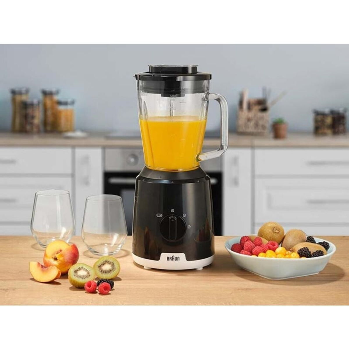 Ein Mixer mit orangener Flüssigkeit steht auf einer weißen Arbeitsplatte. Früchte und Gläser drumherum.