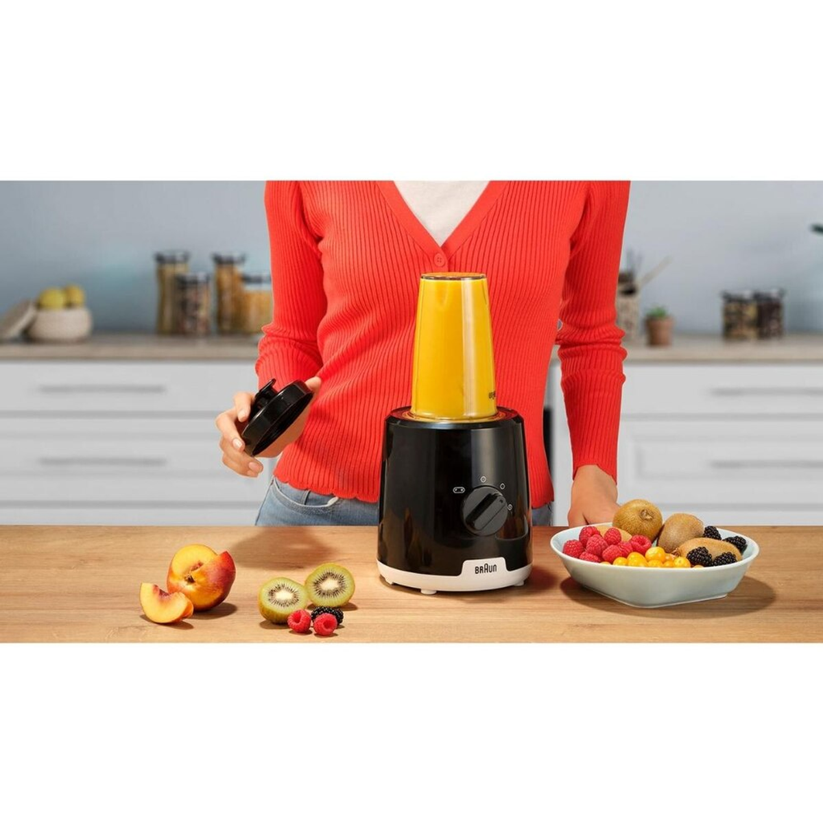 Eine Frau in rotem Hemd in der Nähe eines Mixers, Obst und Mixerteile sichtbar.