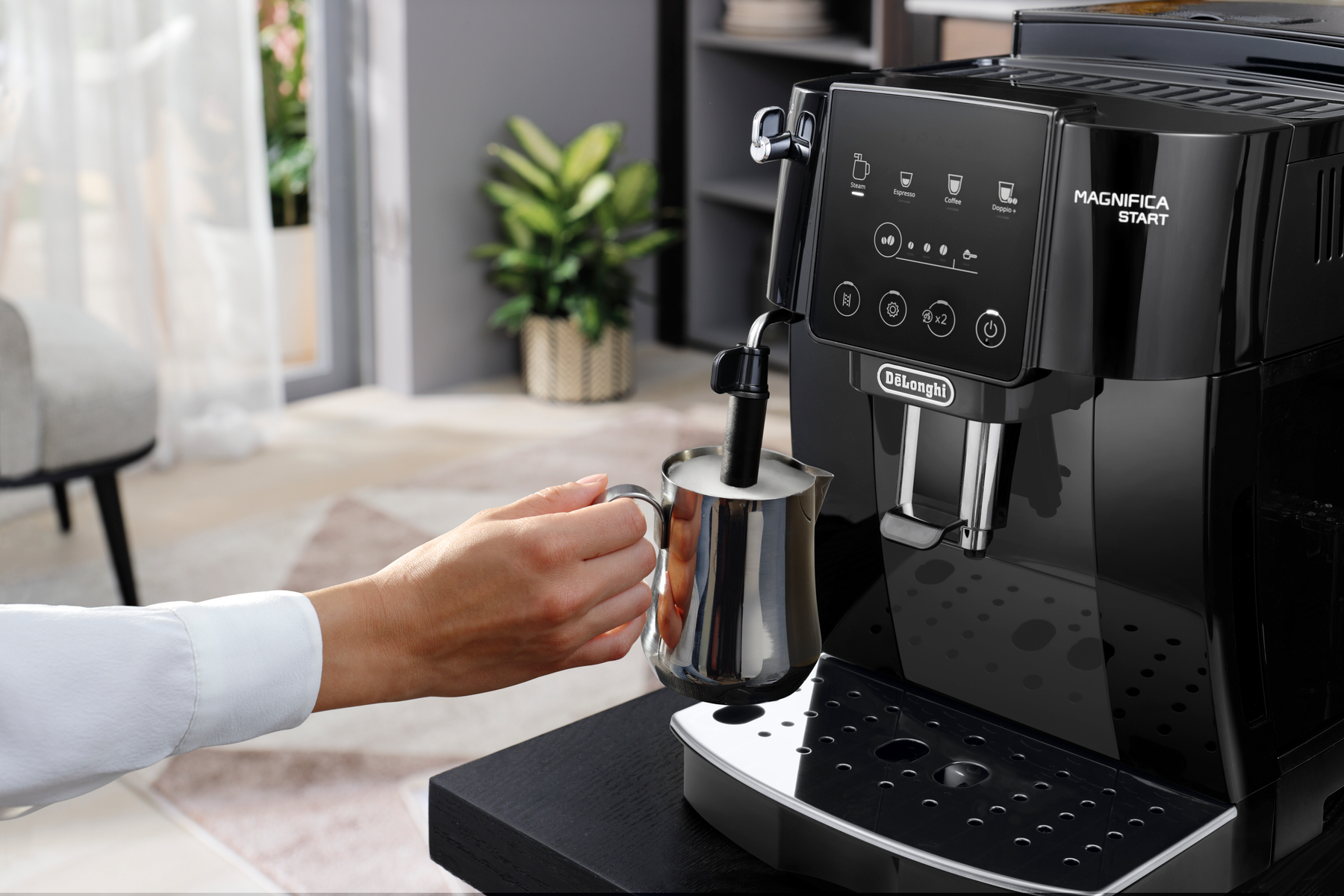 Person gießt Milch in einen Edelstahlkrug neben einer schwarzen DeLonghi Kaffeemaschine.