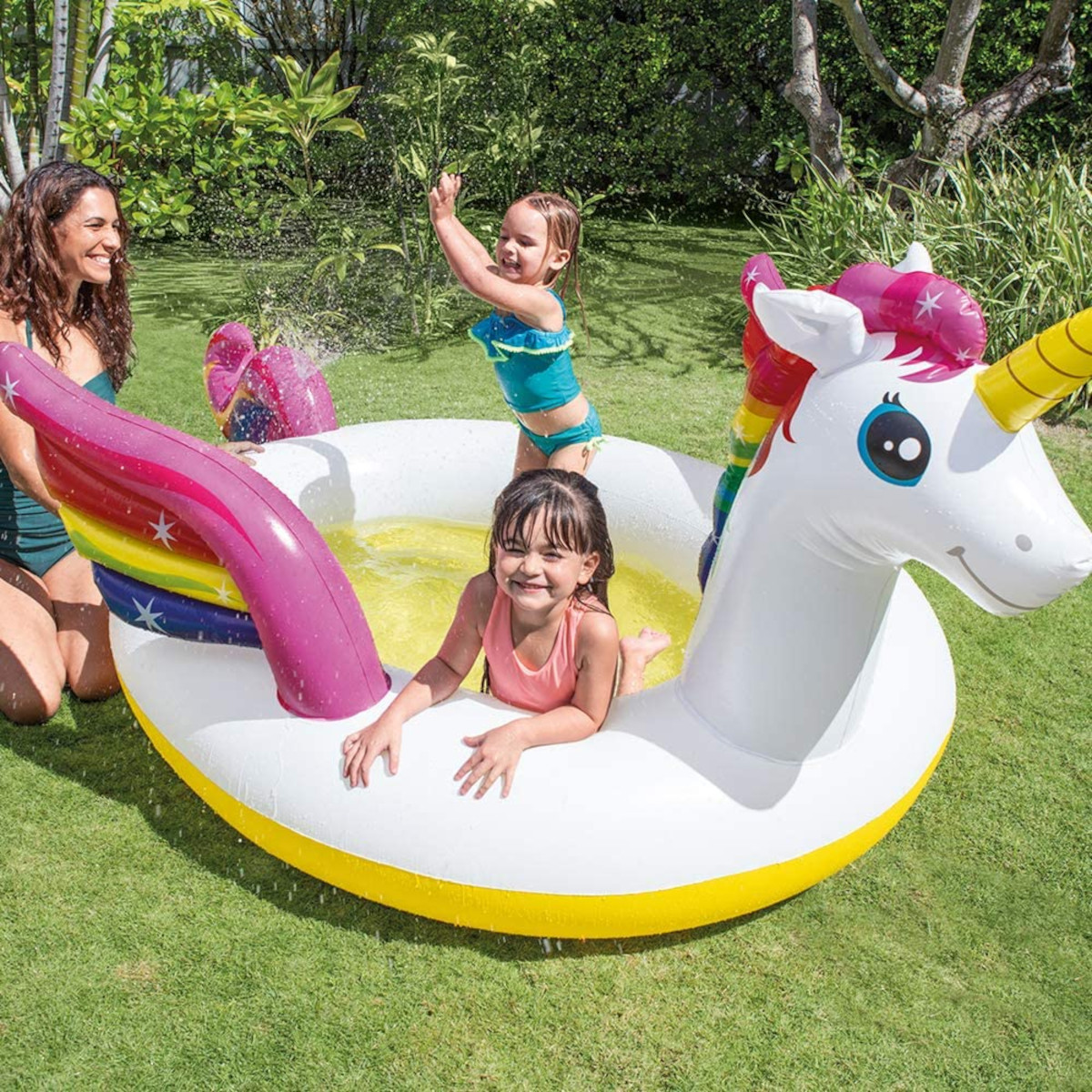 Drei Kinder in einem Einhorn-Pool; eines springt, zwei lächeln. Ein Erwachsener beobachtet. Grüner Rasen im Hintergrund.