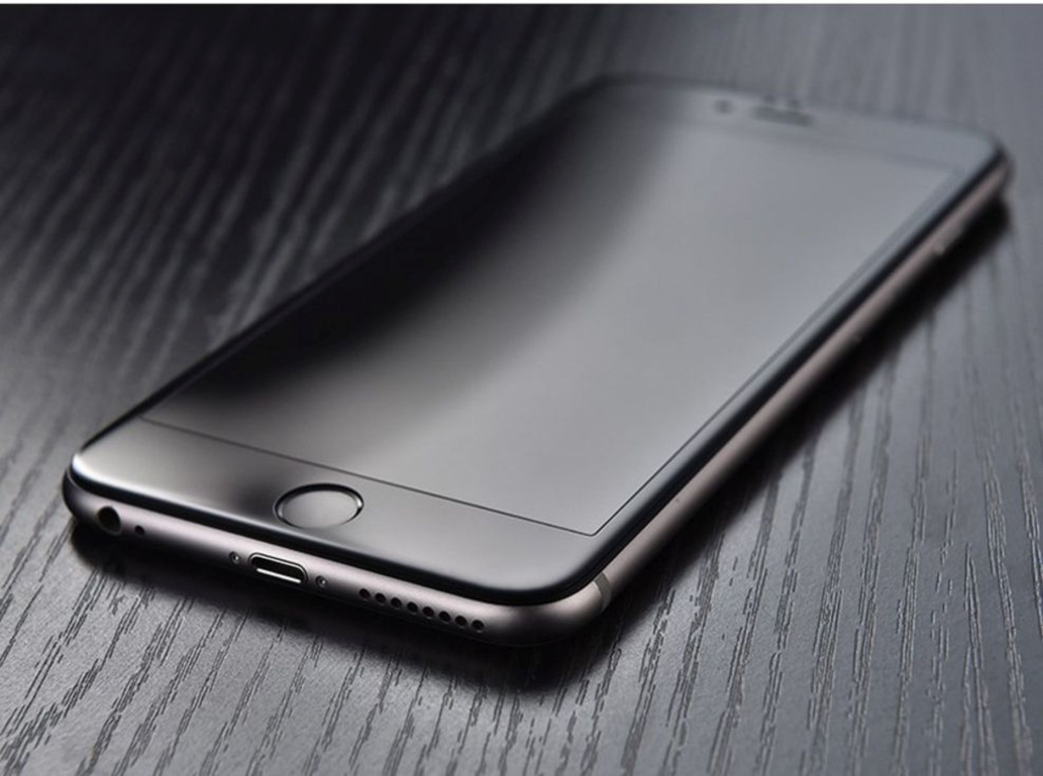 Ein schwarzes Smartphone auf einer Holzoberfläche, mit dunklem Bildschirm, Home-Button und Anschluss.