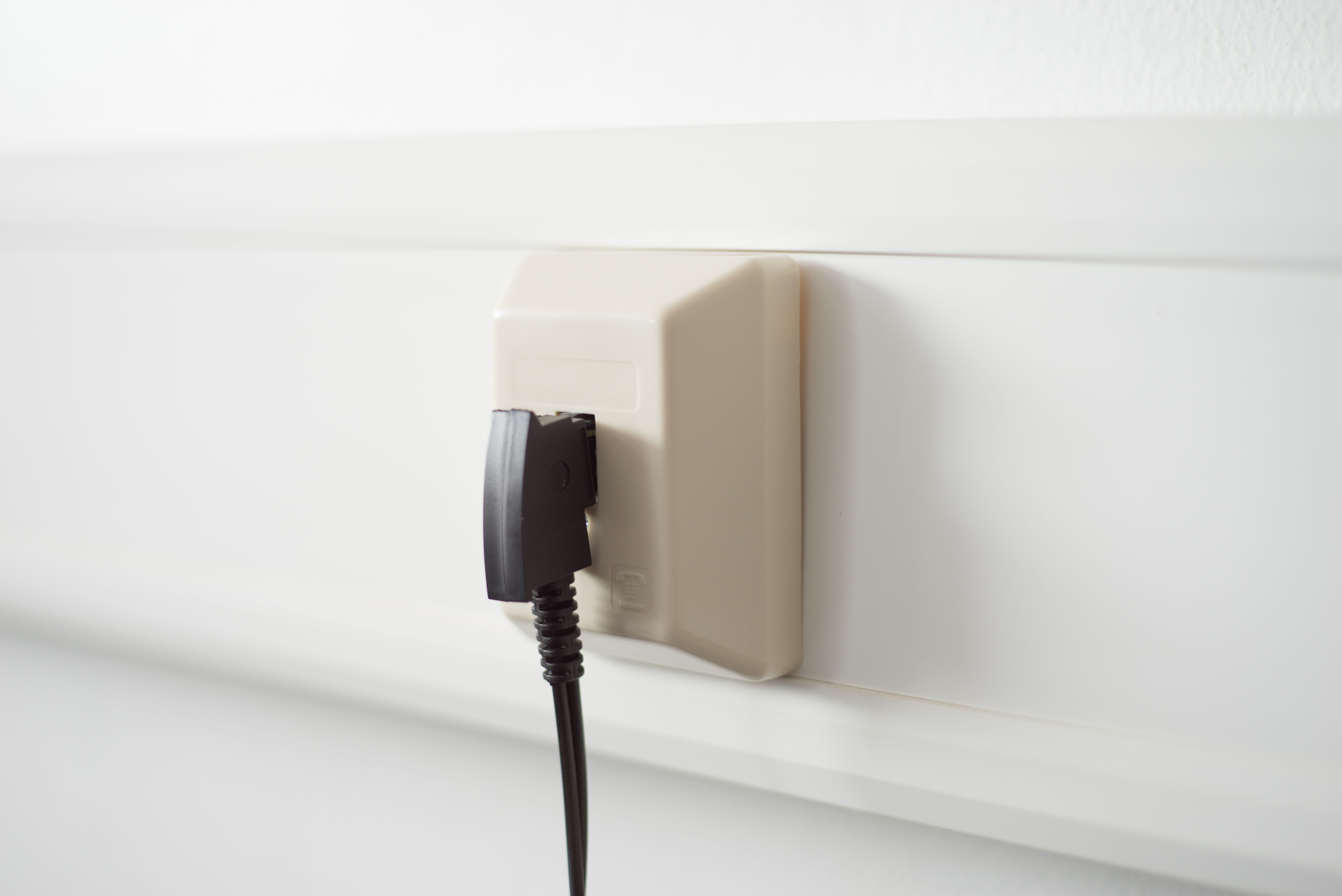 Eine beige Wandsteckdose mit einem eingesteckten schwarzen Stecker, vor einer weißen Wand. Der Stecker steckt halb drin.
