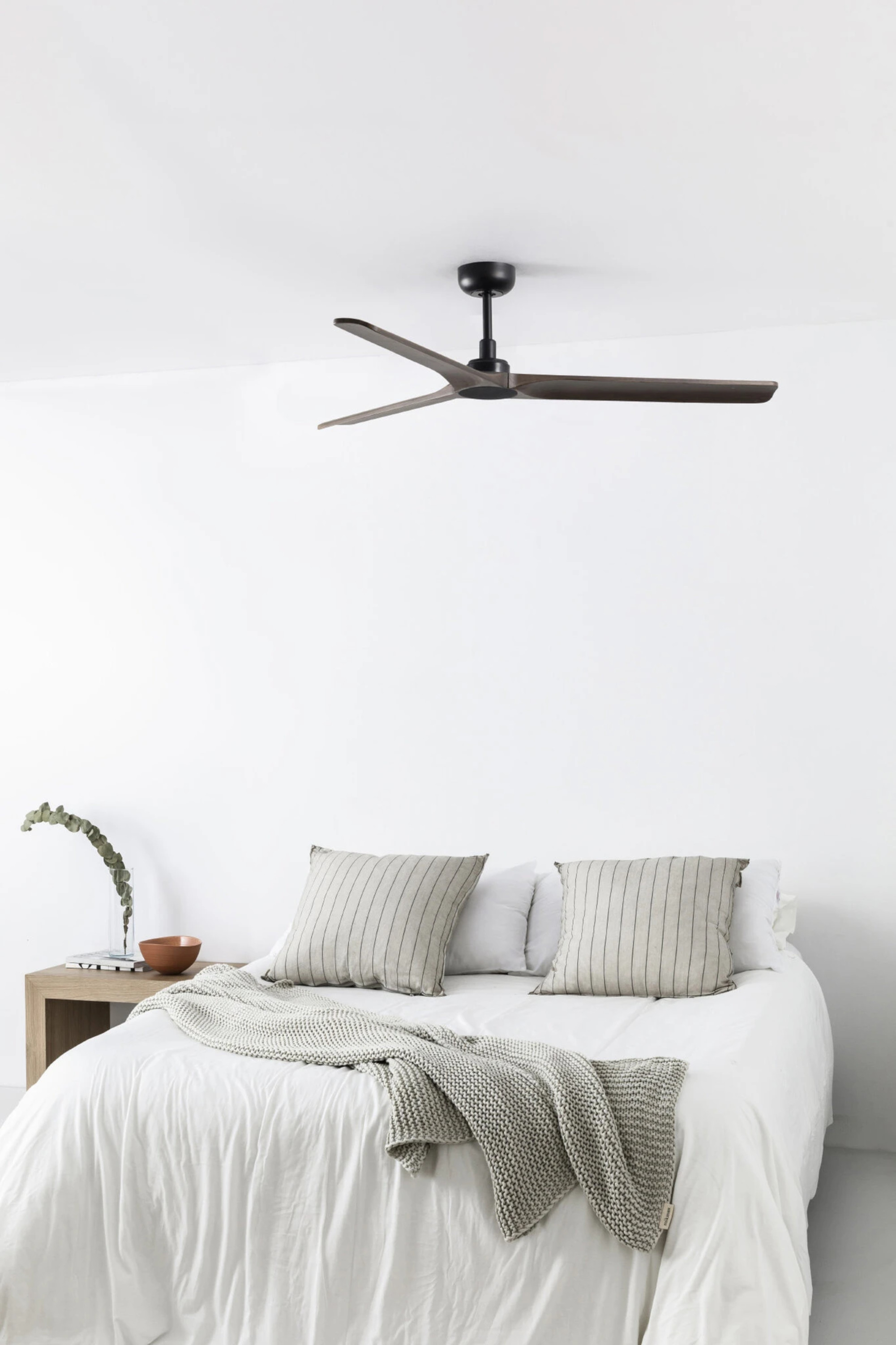 Schlafzimmer mit Deckenventilator, Bett, Kissen und Beistelltisch. Weiße Wände, neutrale Töne, minimalistisches Design.