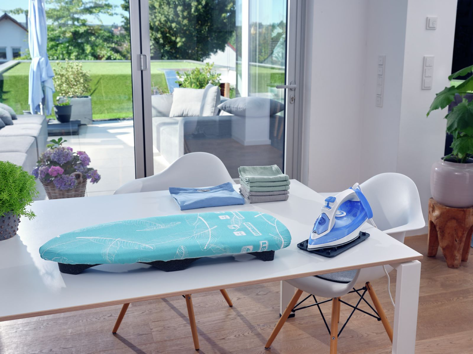 Weißer Tisch mit Bügeleisen, Bügelbrett, gefalteter Kleidung und Gartenblick.