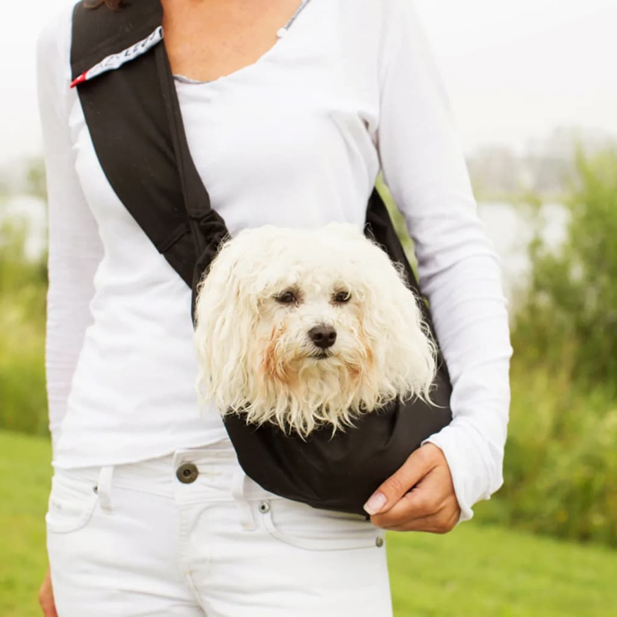 Eine Frau, die einen kleinen weißen Hund in einer schwarzen Sling-Tasche trägt, lächelnd.