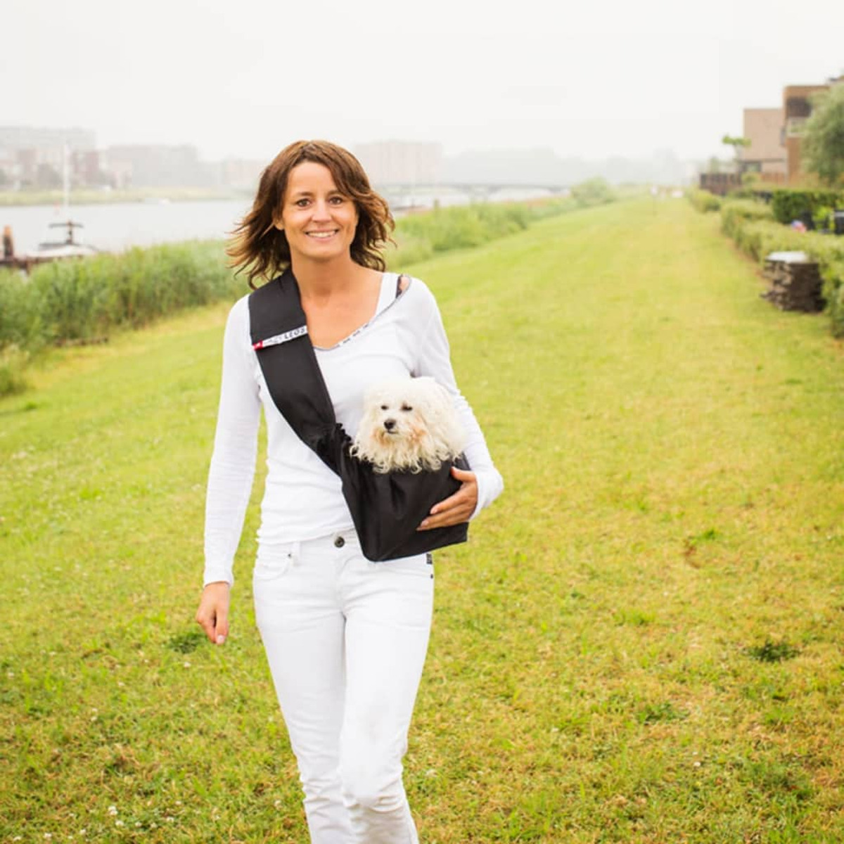 Frau geht mit Hund in einer Tragetasche spazieren. Sie befinden sich in einer parkähnlichen Umgebung.