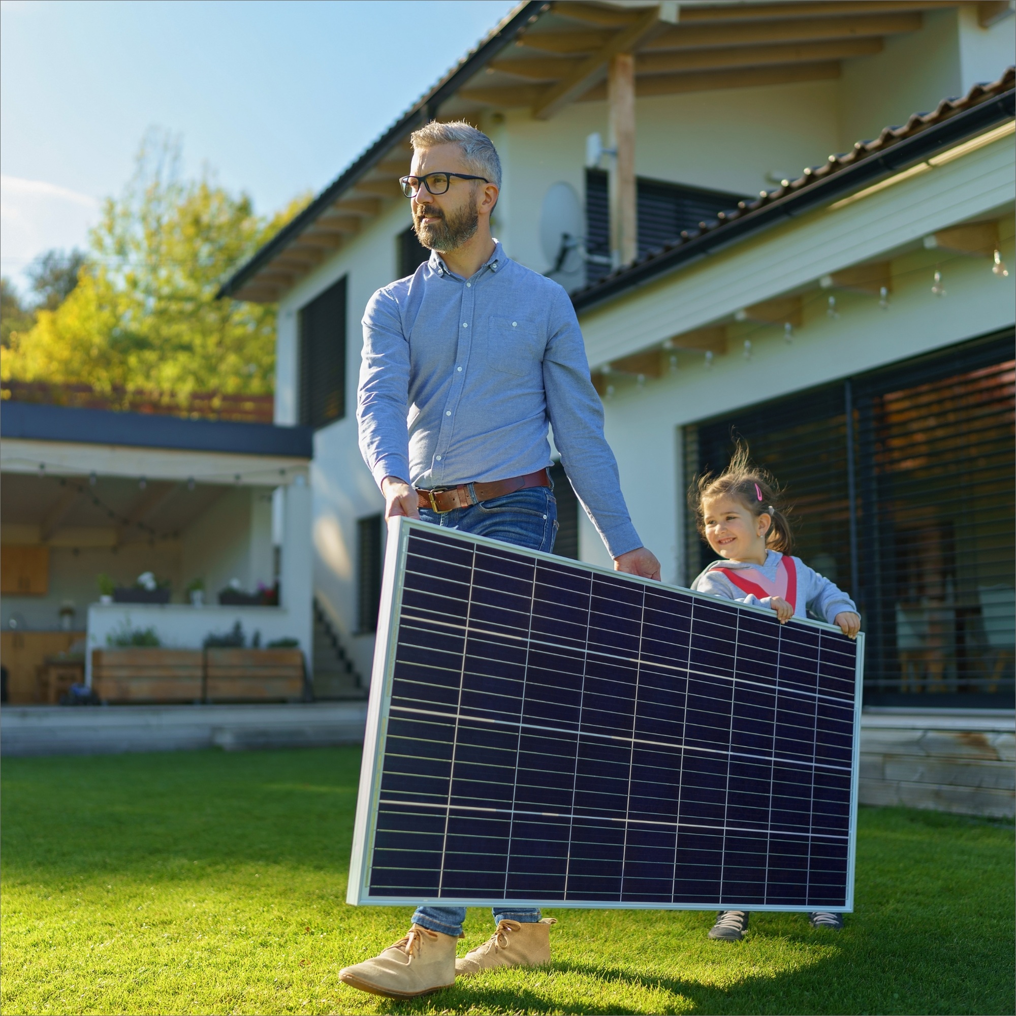 Ein Mann und ein Kind tragen ein Solarpanel im Garten, Haus im Hintergrund.