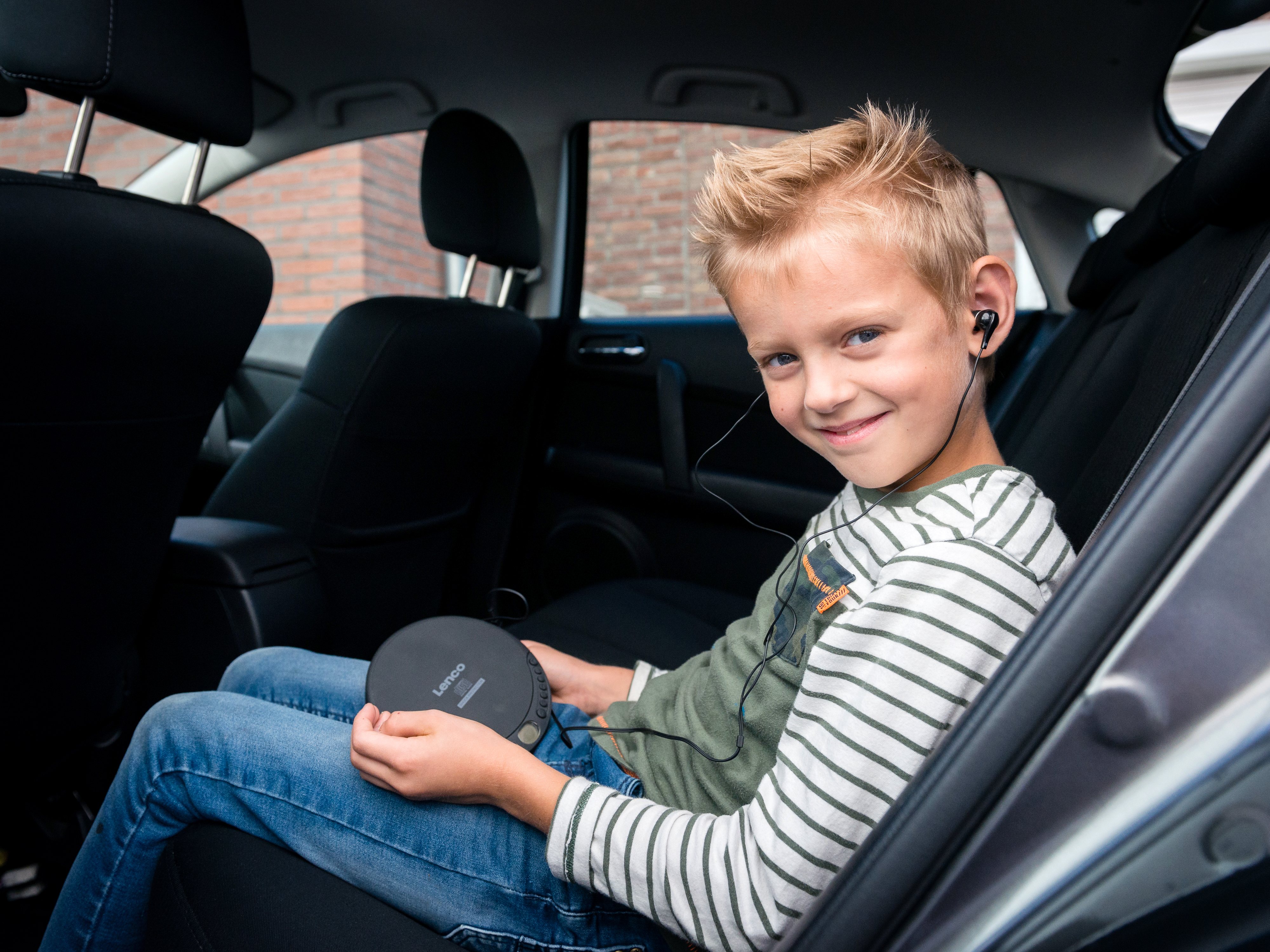 Uśmiechnięty chłopiec w słuchawkach siedzi na tylnym siedzeniu samochodu, trzymając odtwarzacz CD.