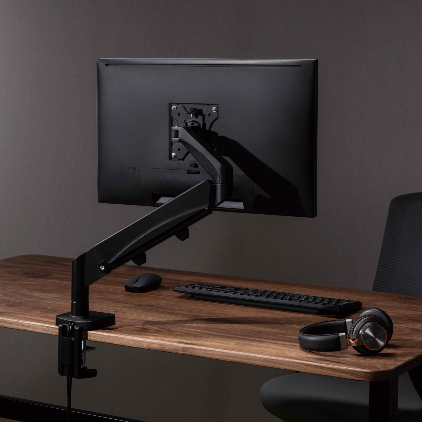 Schwarzer Monitorarm, Monitor, Tastatur, Maus und Kopfhörer auf einem Holzschreibtisch. Dunkler Hintergrund.
