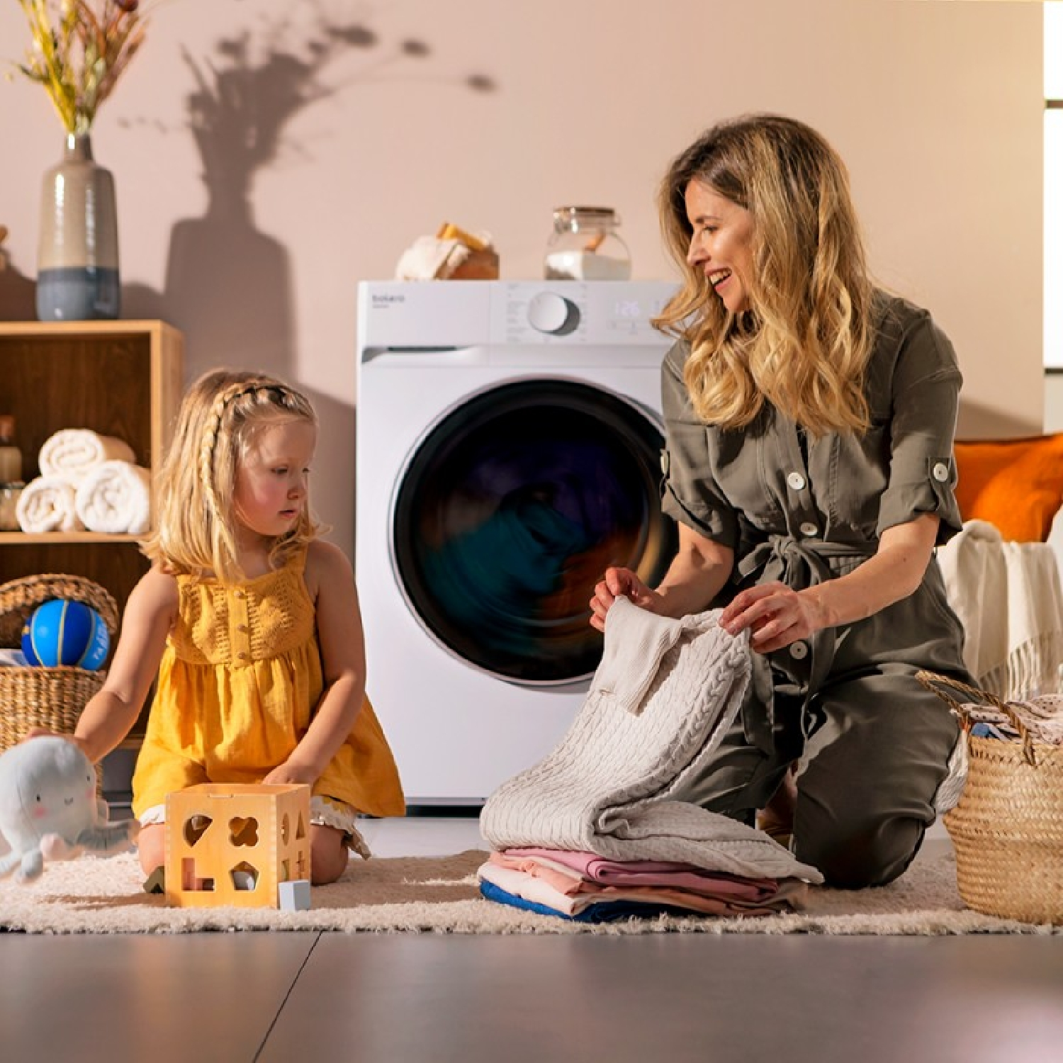 Kobieta i dziecko składają pranie obok pralki, dywanu i zabawki.