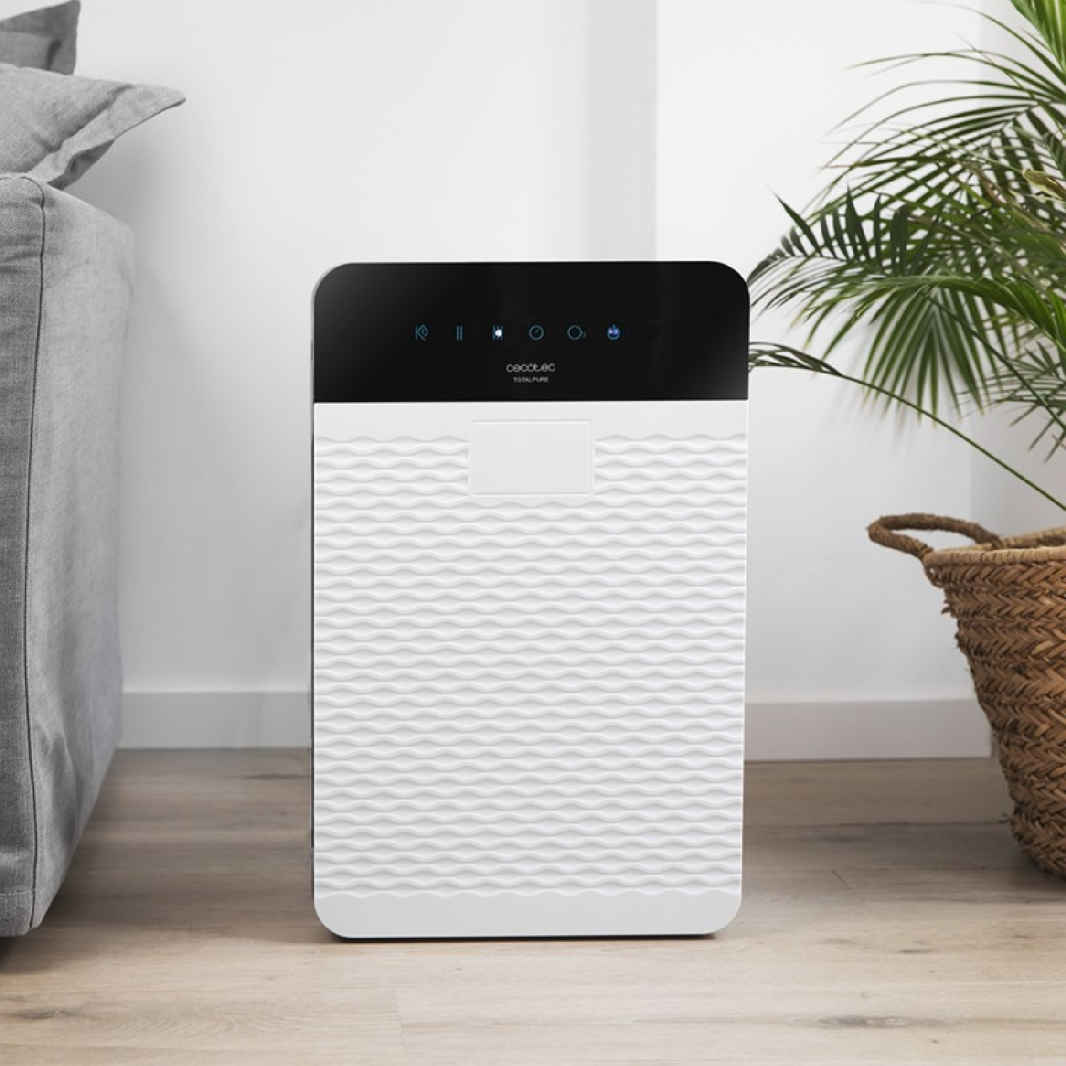 Purificador de aire blanco con un panel frontal texturizado ondulado. Una planta y un sofá gris en el fondo.