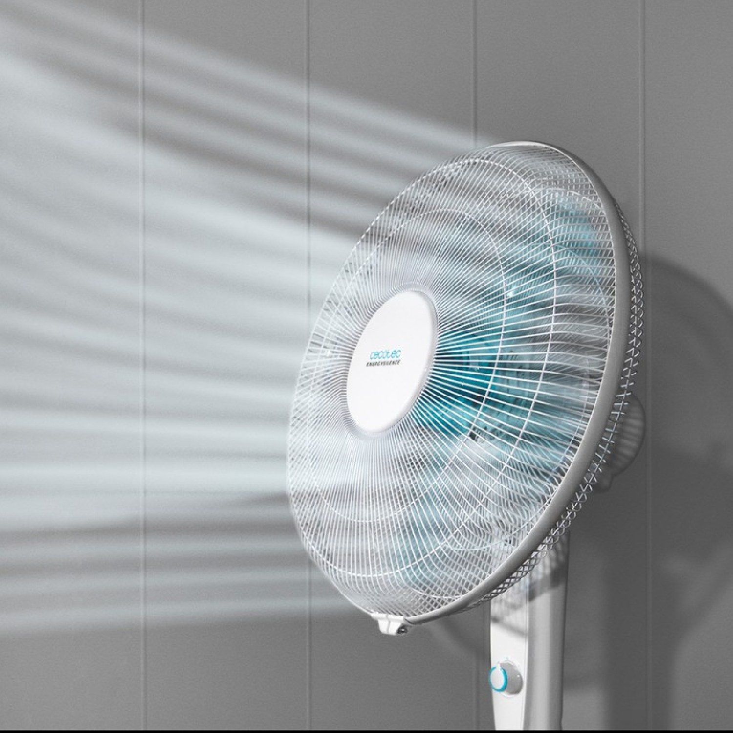 Ventilador blanco con aspas azules, inclinado a la derecha, contra una pared gris.