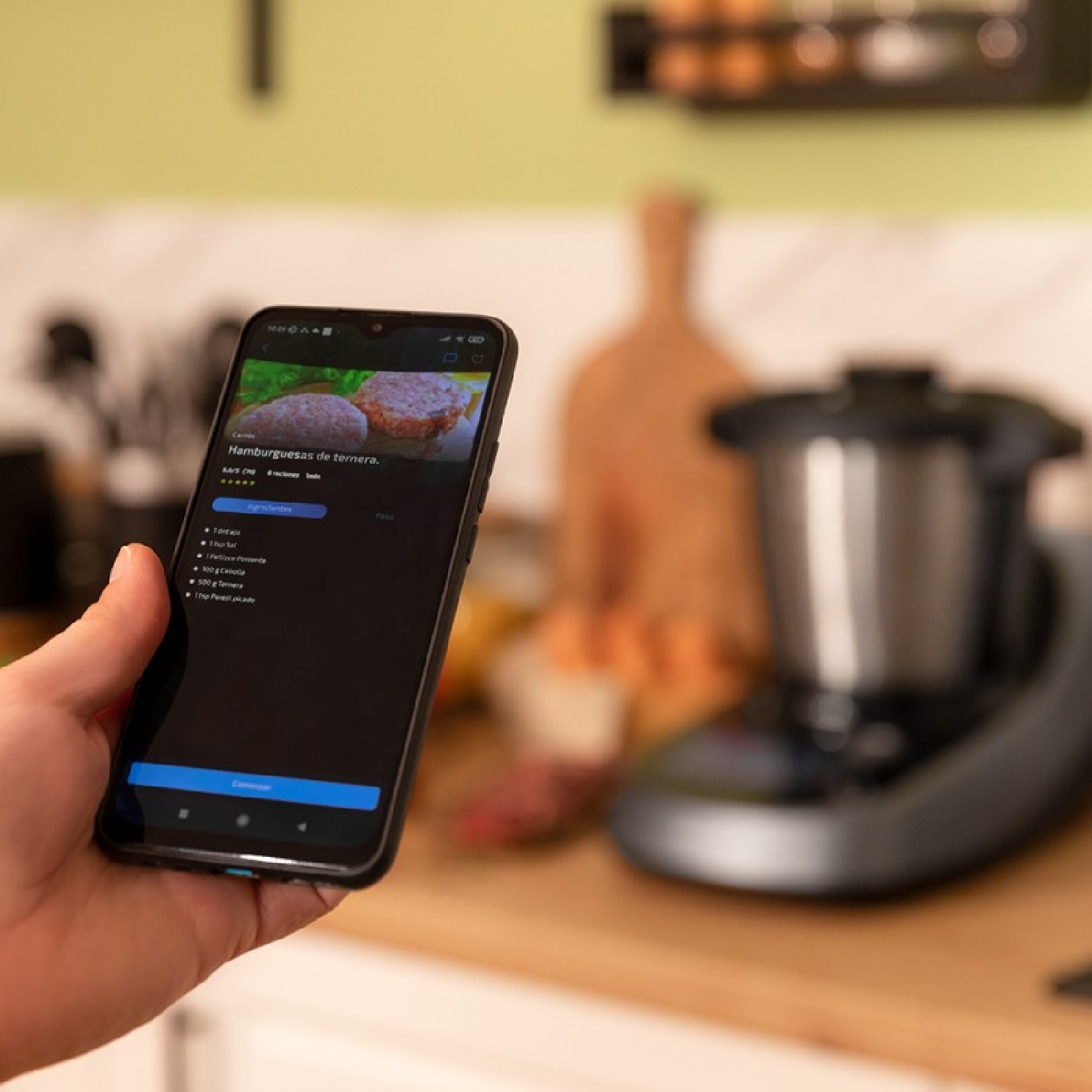 Una persona tiene in mano uno smartphone con una ricetta per hamburger. Un elettrodomestico da cucina è sfocato.