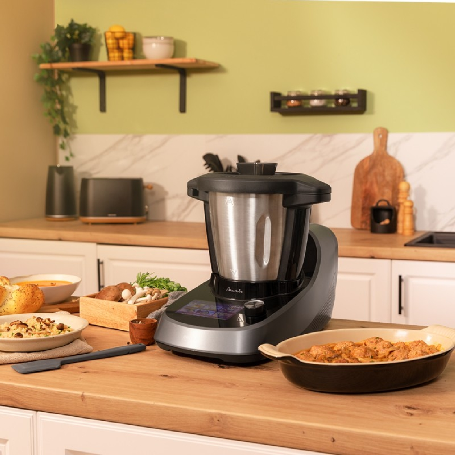 Una cucina con un robot da cucina, piatti e ingredienti su un bancone di legno. Parete verde.