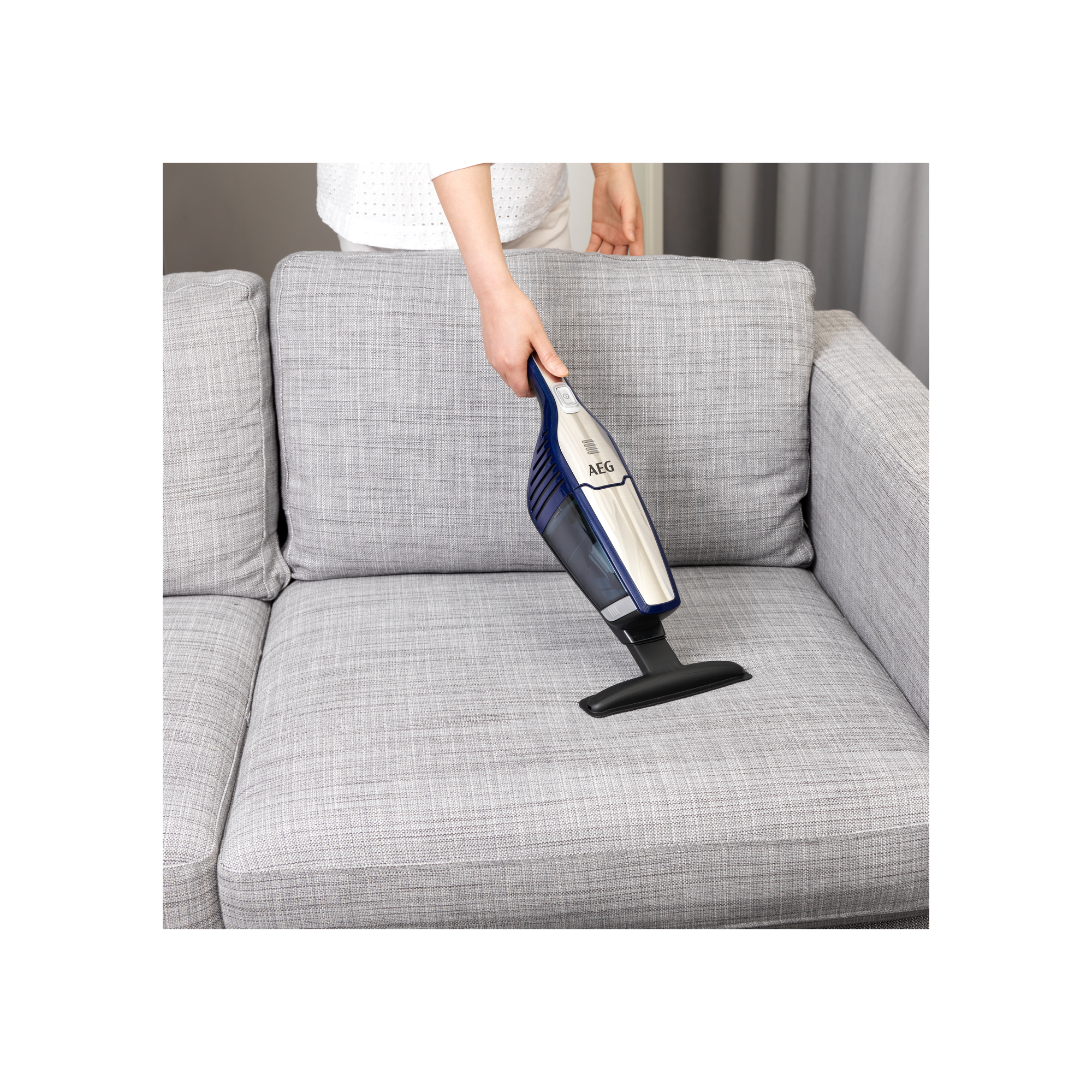 Osoba używająca niebiesko-białego odkurzacza ręcznego AEG do czyszczenia szarej sofy.
